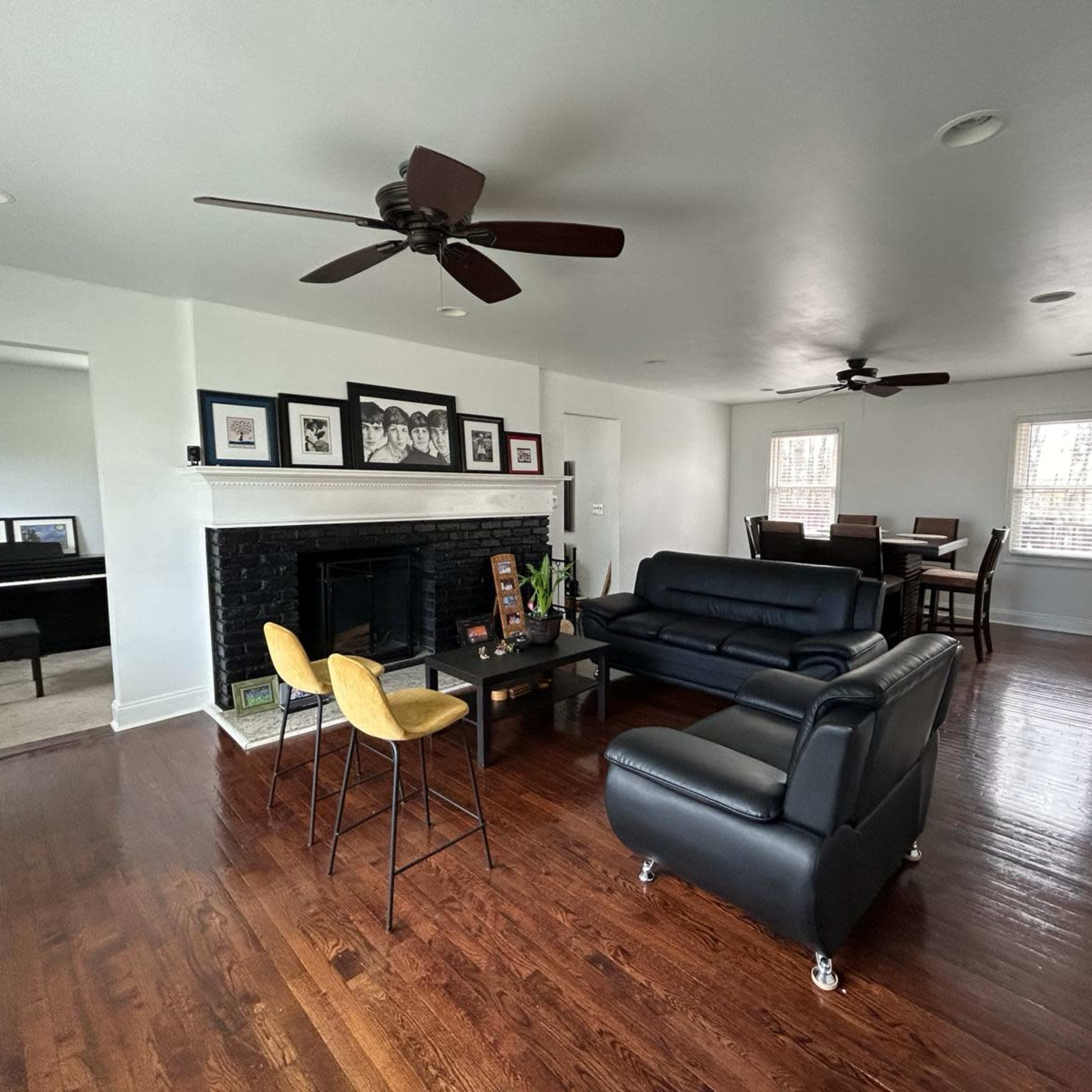 The living room features a black leather sofa, a coffee table, and a dining area, with a piano visible in the background and two ceiling fans overhead.