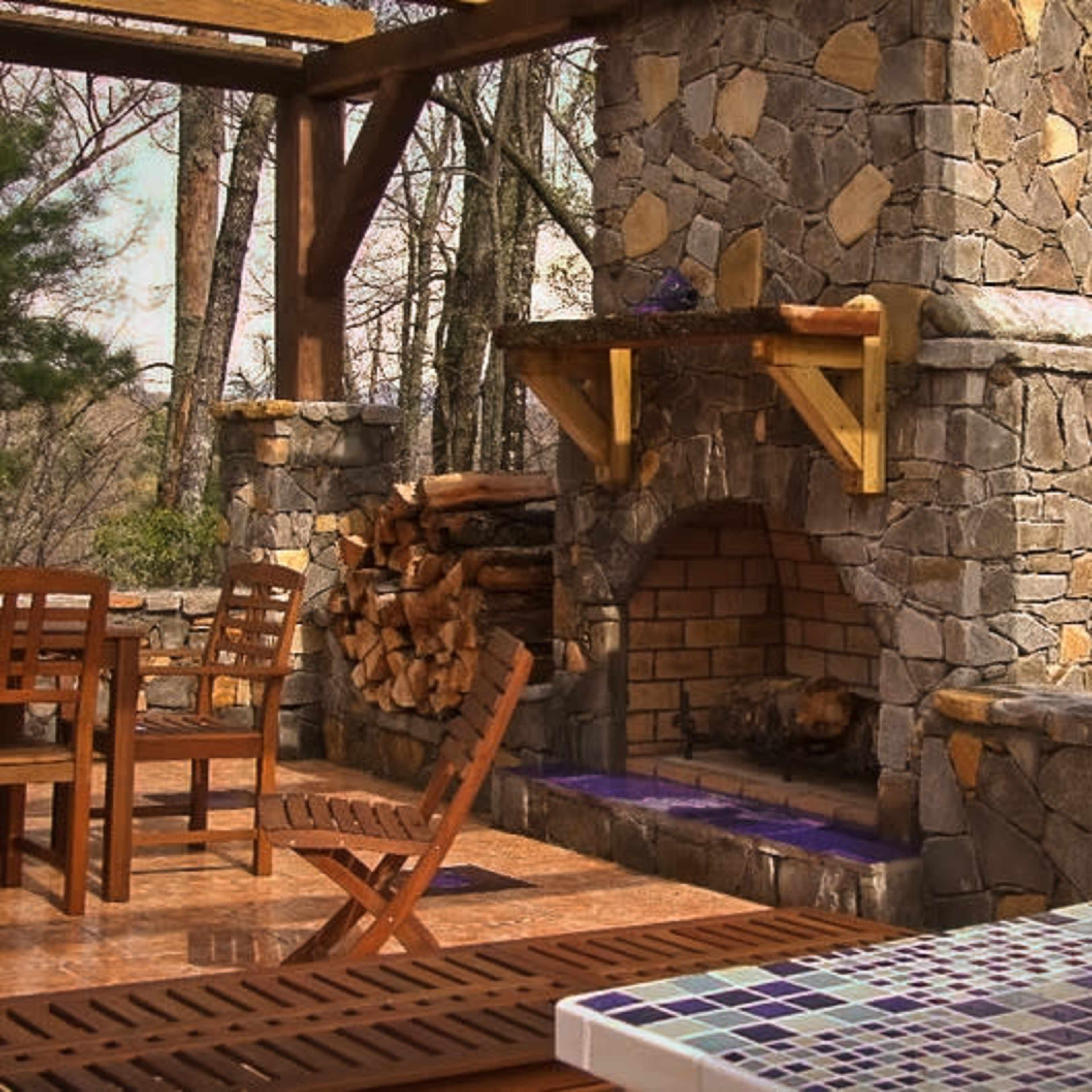 An outdoor stone fireplace with a woodpile, set on a wooden deck surrounded by trees.