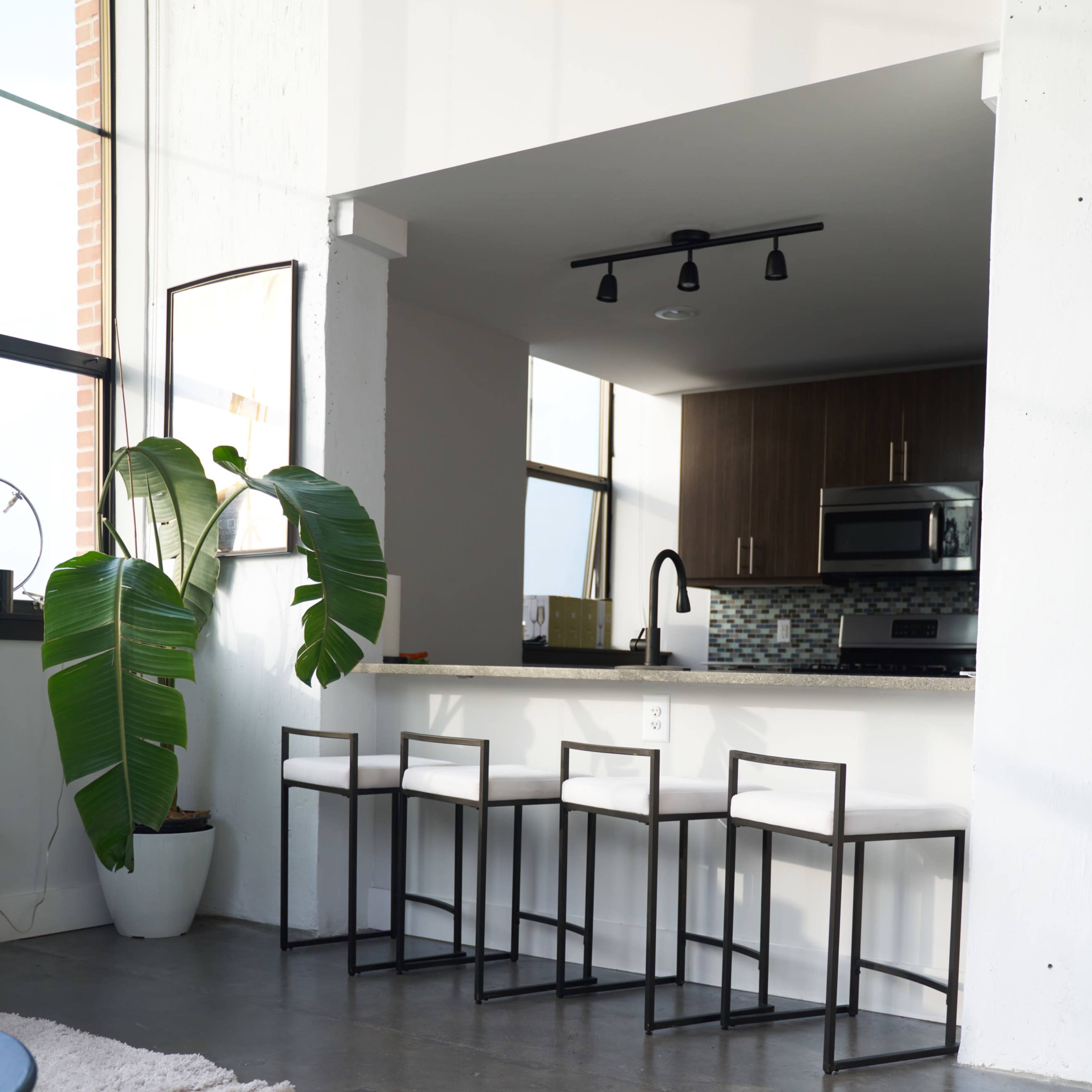 A spacious kitchen area features a row of four bar stools at a counter, with large windows and a potted plant in the background.