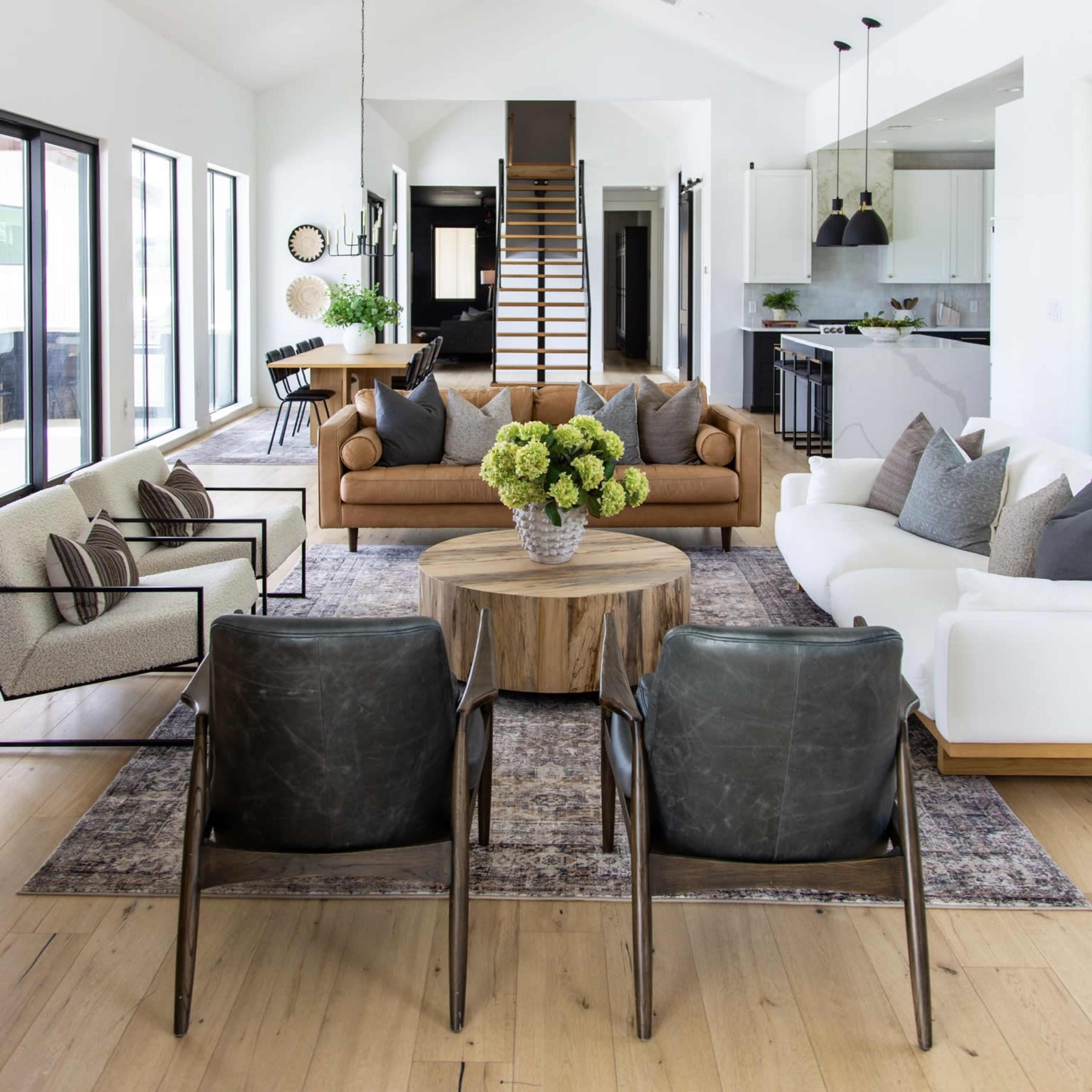 A modern living room with a brown sofa, two black chairs, a round wooden coffee table, and large windows overlooking a staircase and dining area.