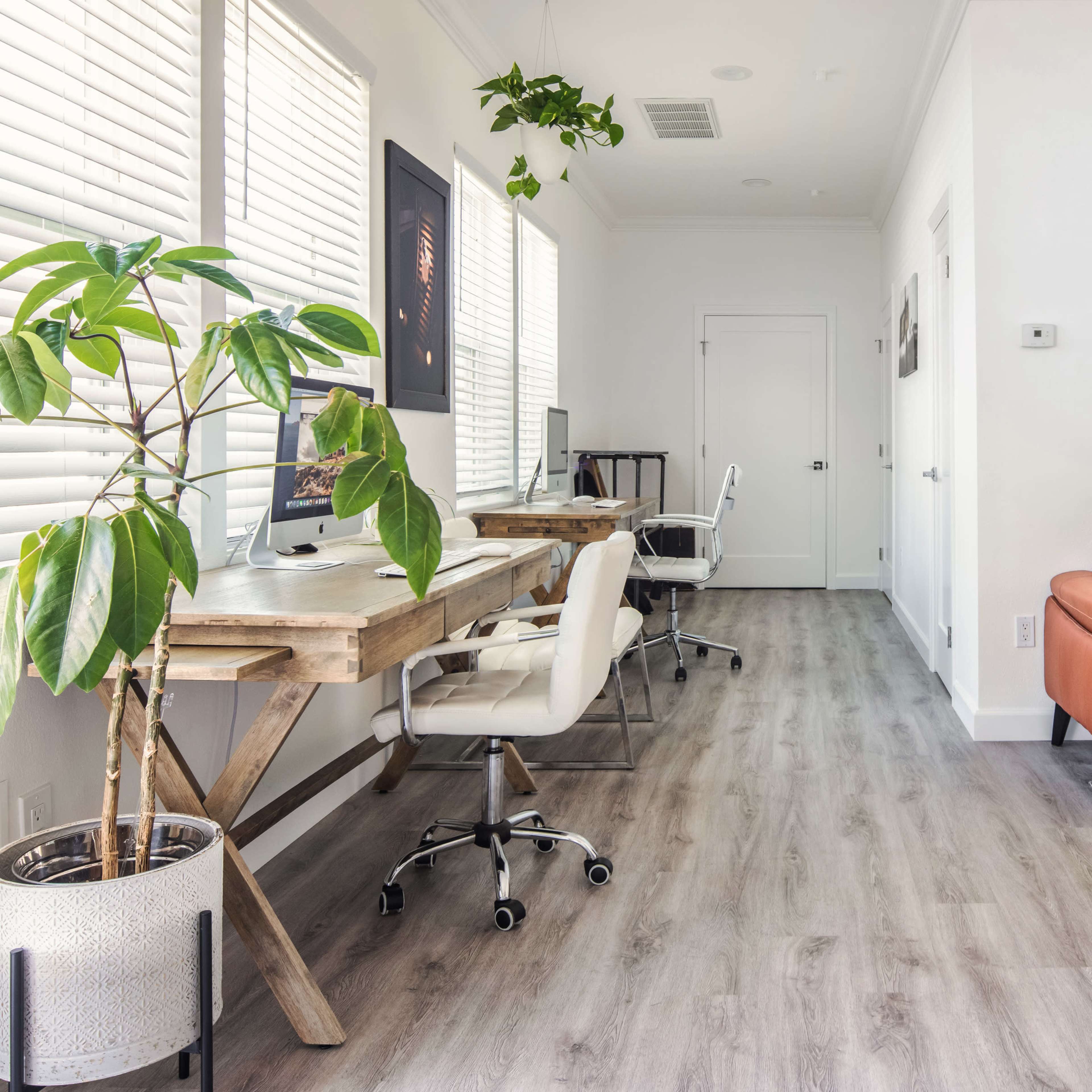 A modern interior office space features two work desks, a brown leather sofa, and potted plants, with ample natural light from the window.