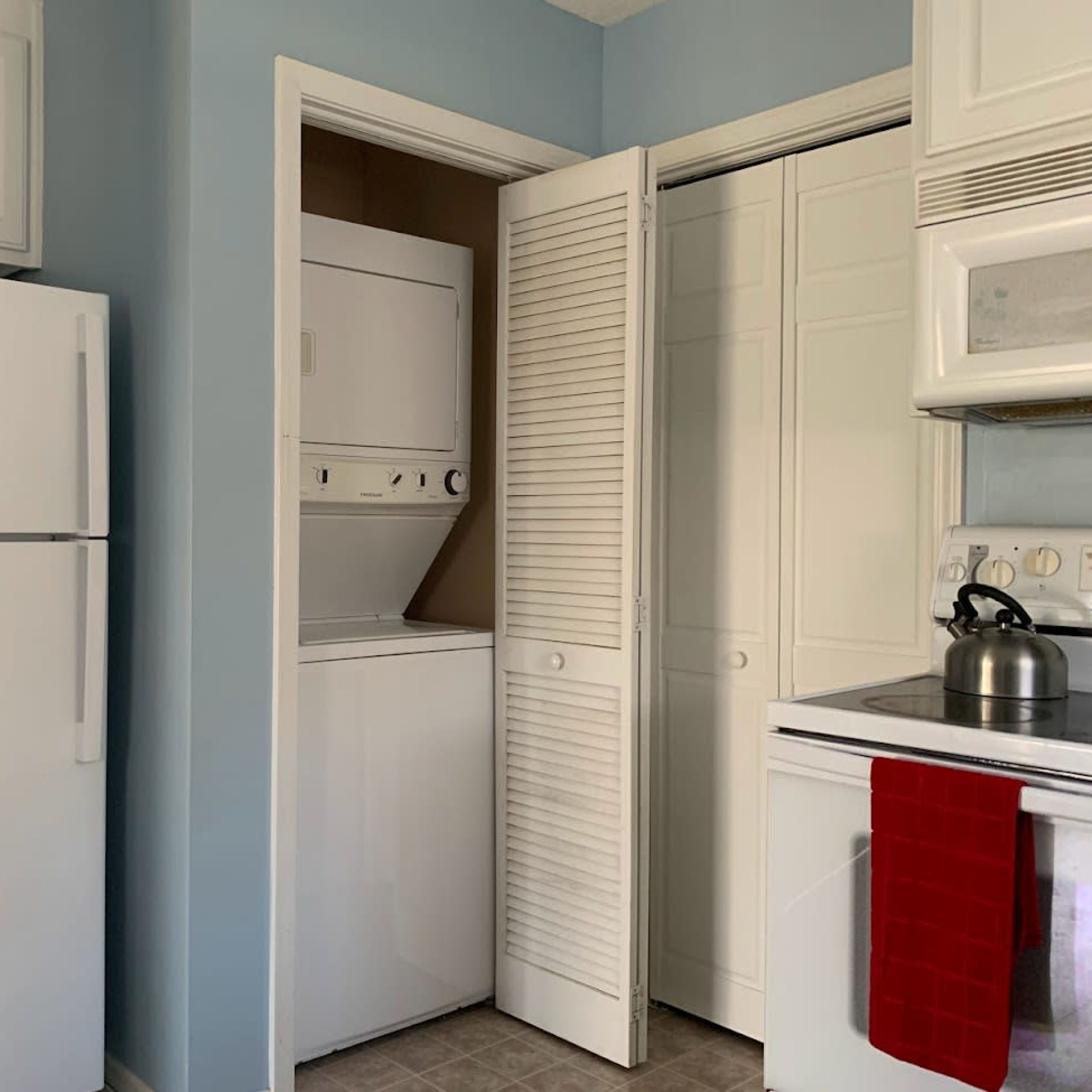 A kitchen featuring a refrigerator, a stacked washing machine and dryer, and a stove with a kettle on it, all set against light blue walls.