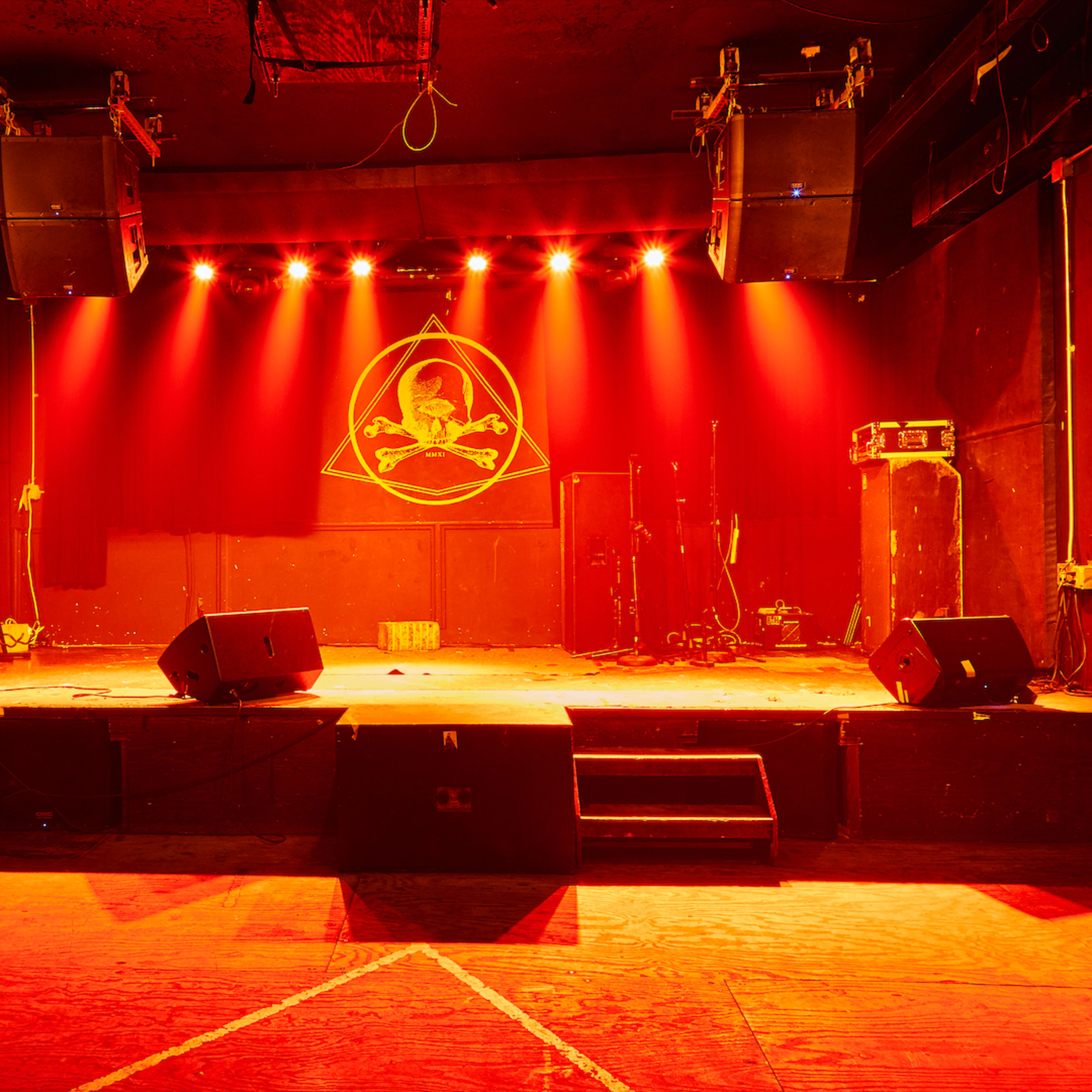 A dimly lit stage with a red hue, featuring a circular logo on the backdrop and audio equipment positioned on either side.
