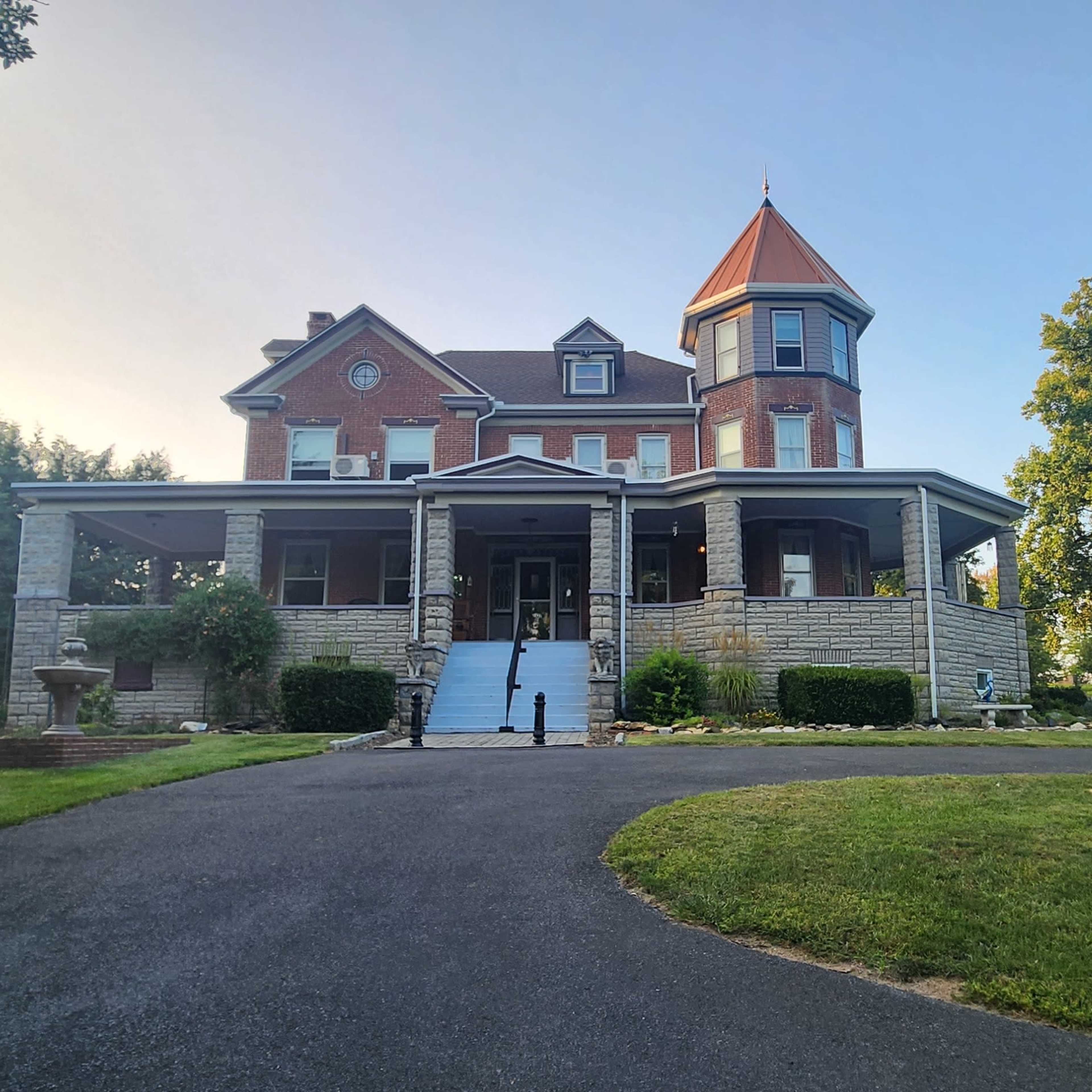 A large, historic house with a turret and a wrap-around porch is surrounded by well-maintained lawns and trees.