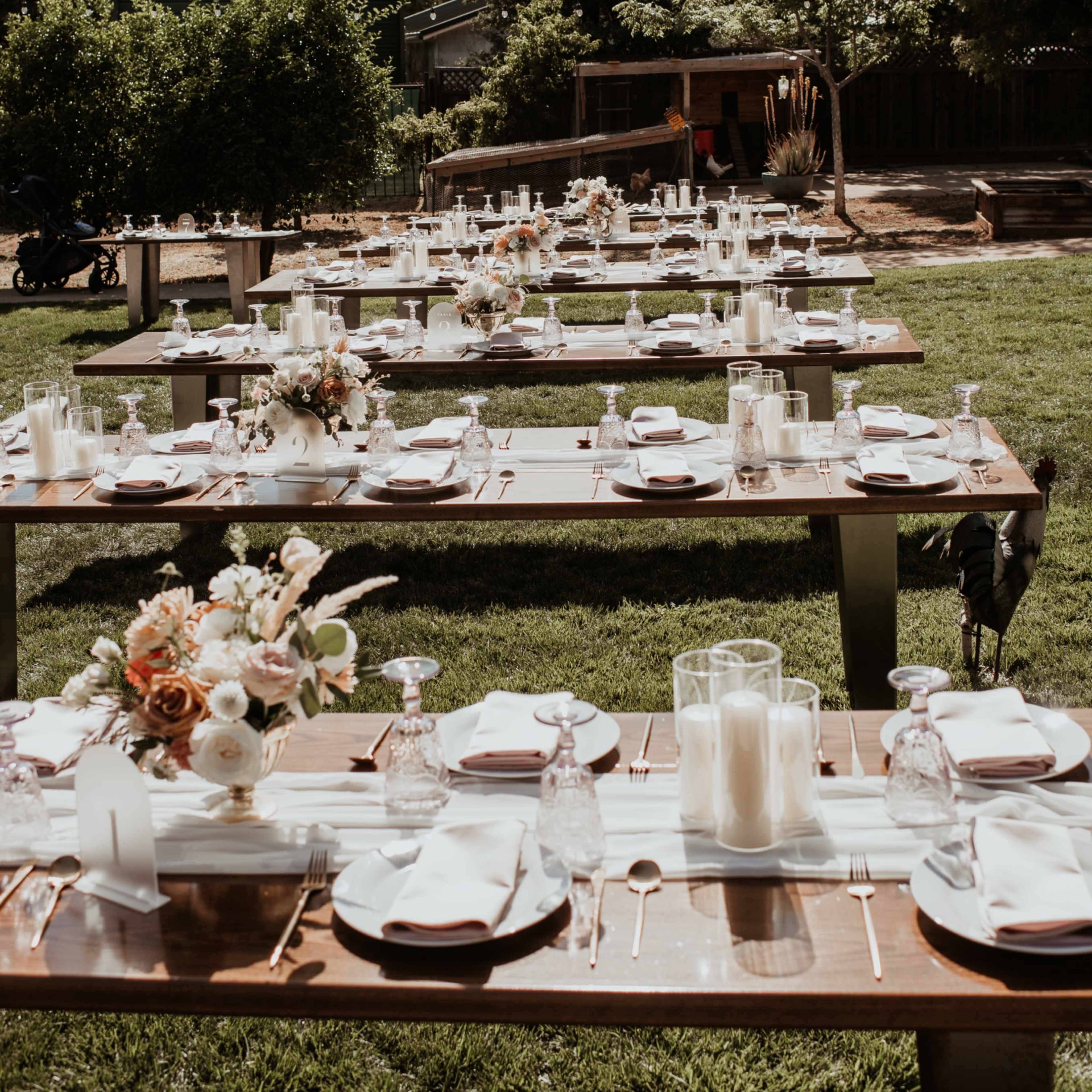 A long outdoor dining setup features multiple wooden tables adorned with elegant place settings and floral centerpieces in a grassy area.