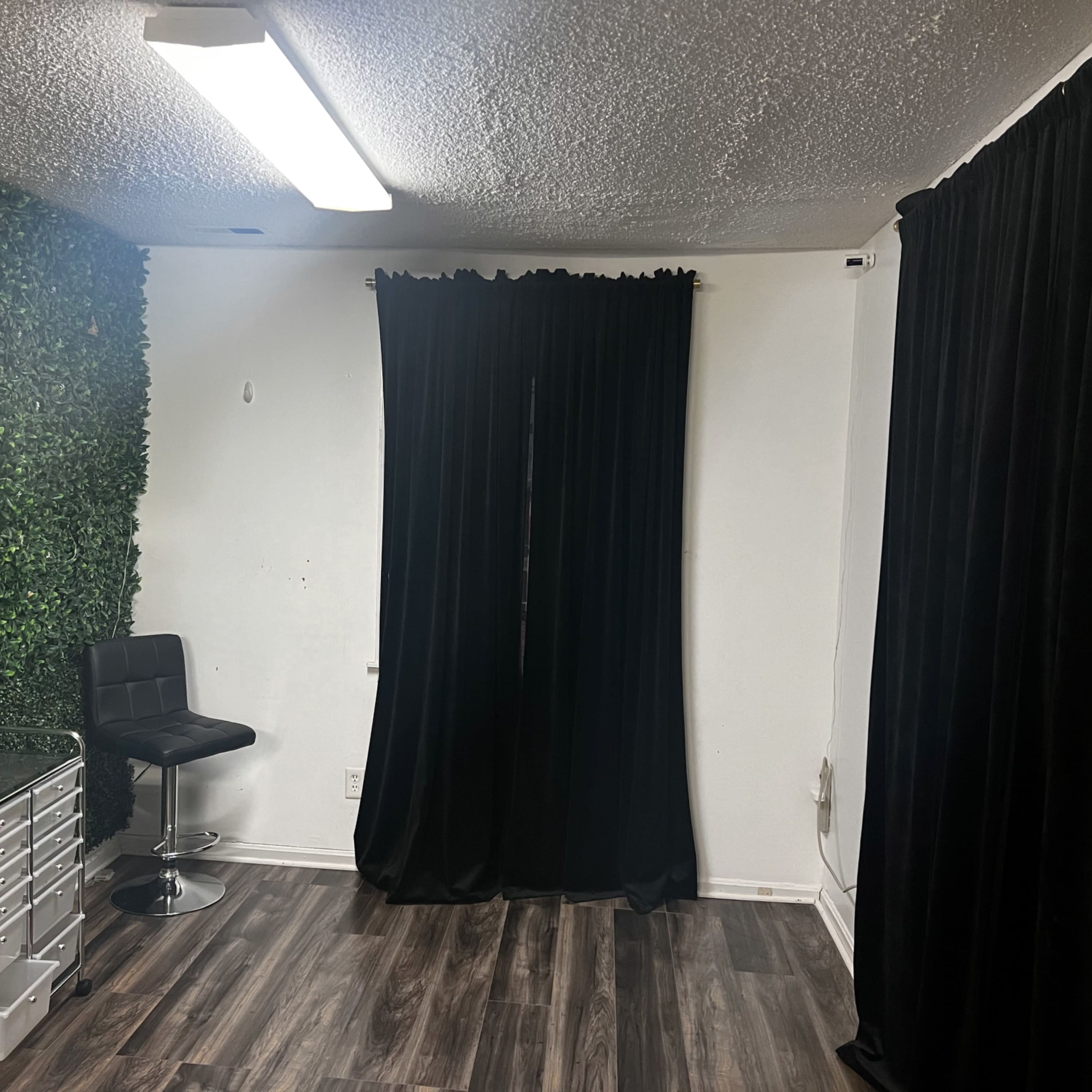 A room with a green plant wall, a black chair, a storage cart, and black curtains covering a window.