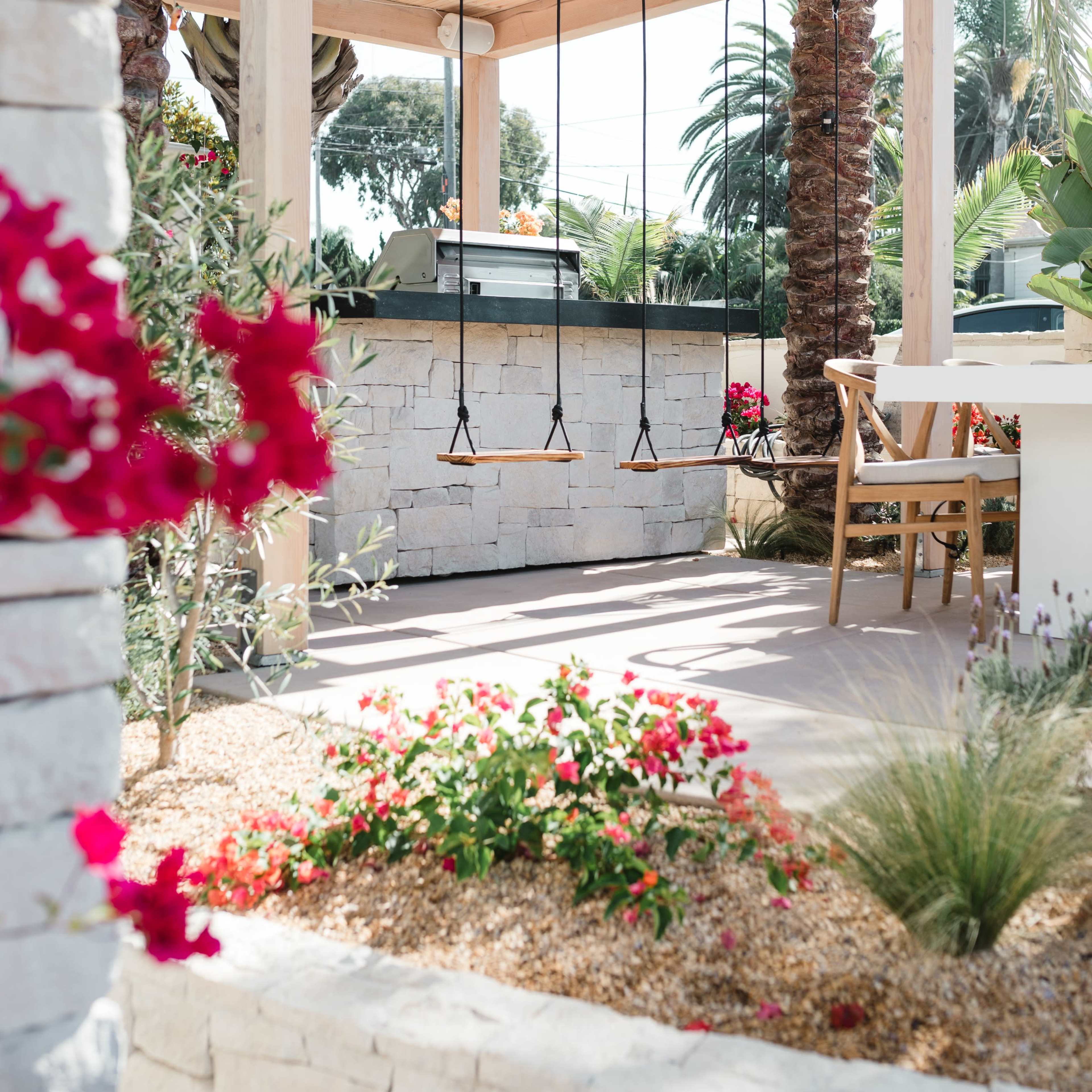 A wooden pergola with hanging swings is set beside a stone grill area, surrounded by colorful flowers and palm trees.