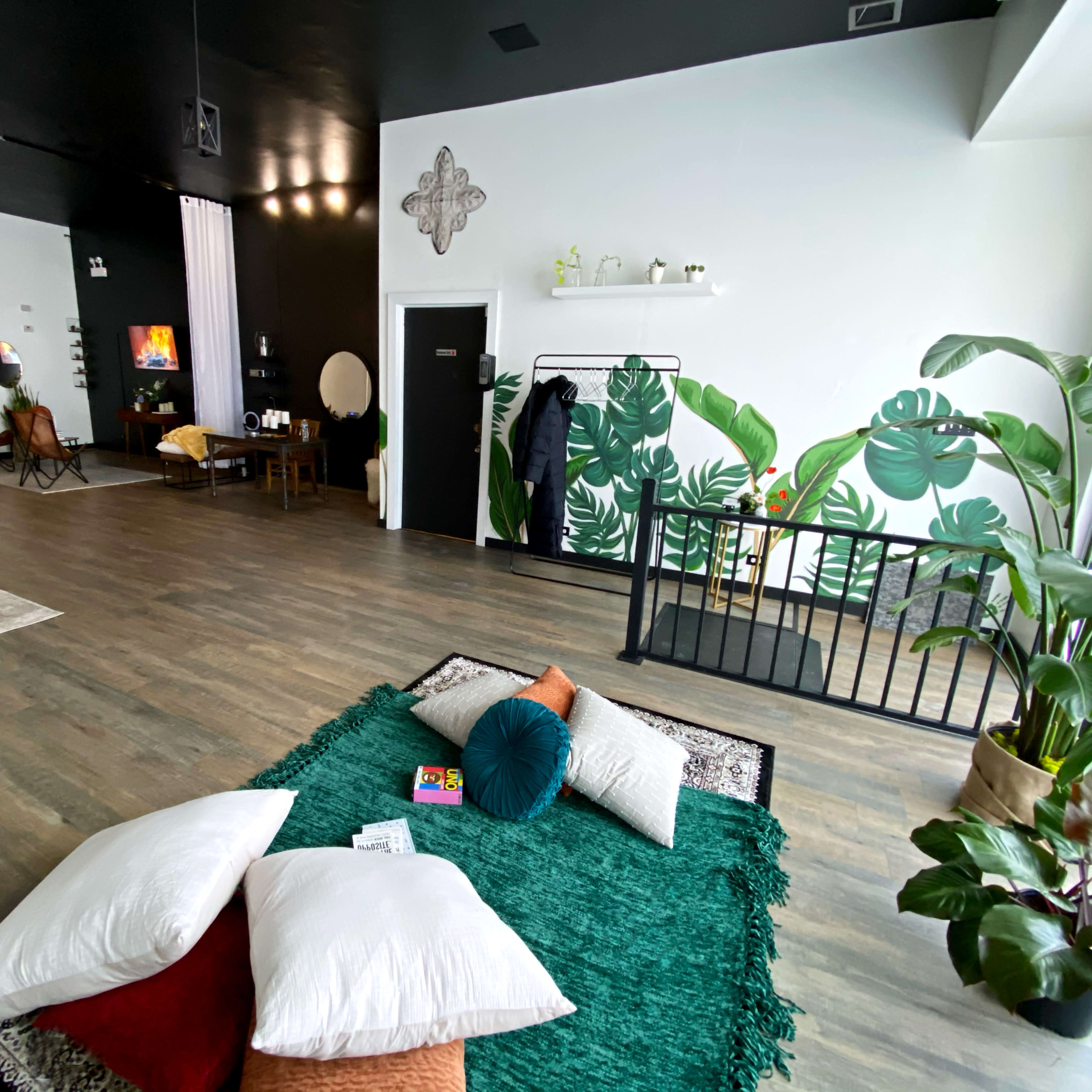 A spacious interior with a green rug and scattered cushions, accented by large plants and a wall featuring a tropical mural.