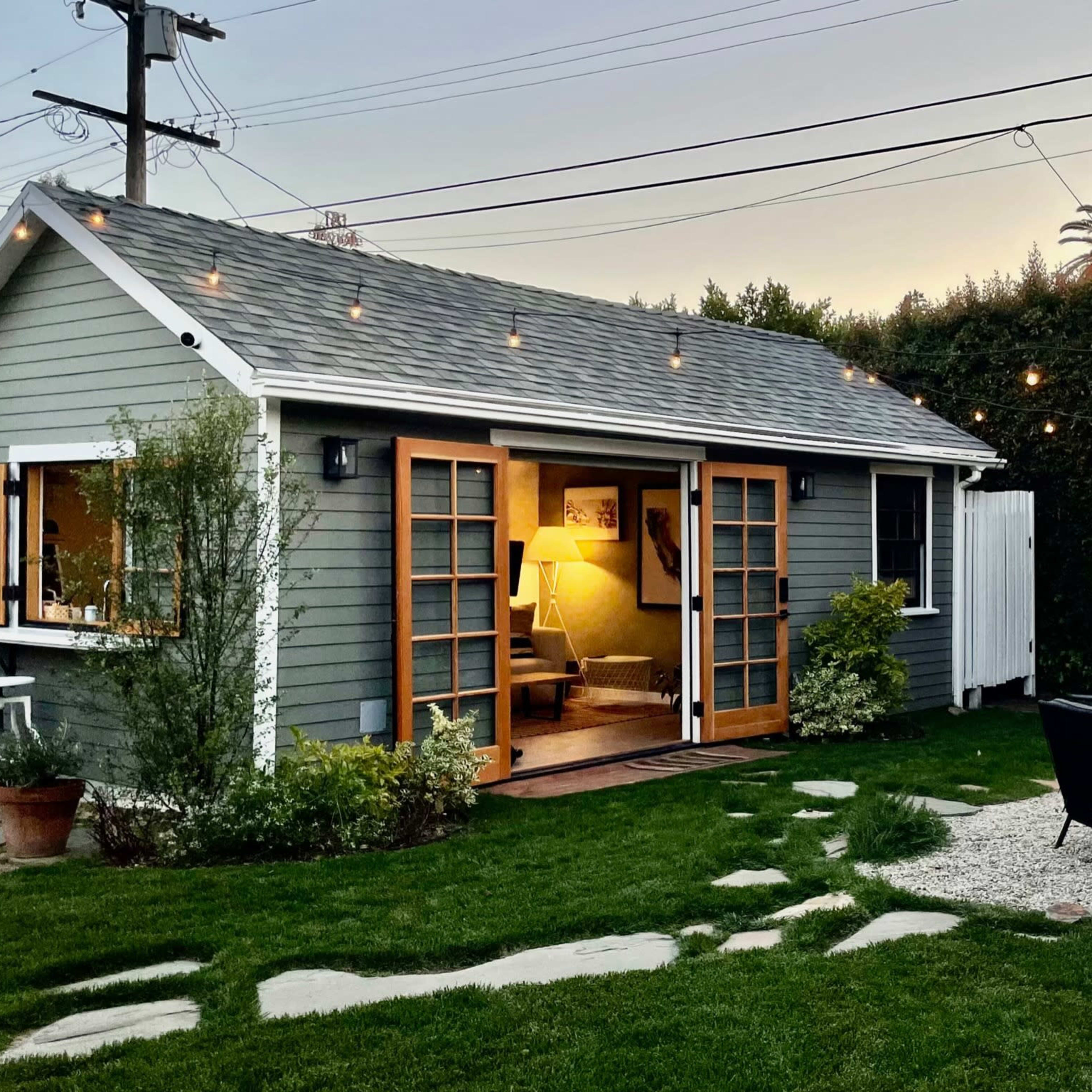A cozy cottage with French doors opens to a backyard featuring a grassy area, outdoor seating, and string lights among the greenery.