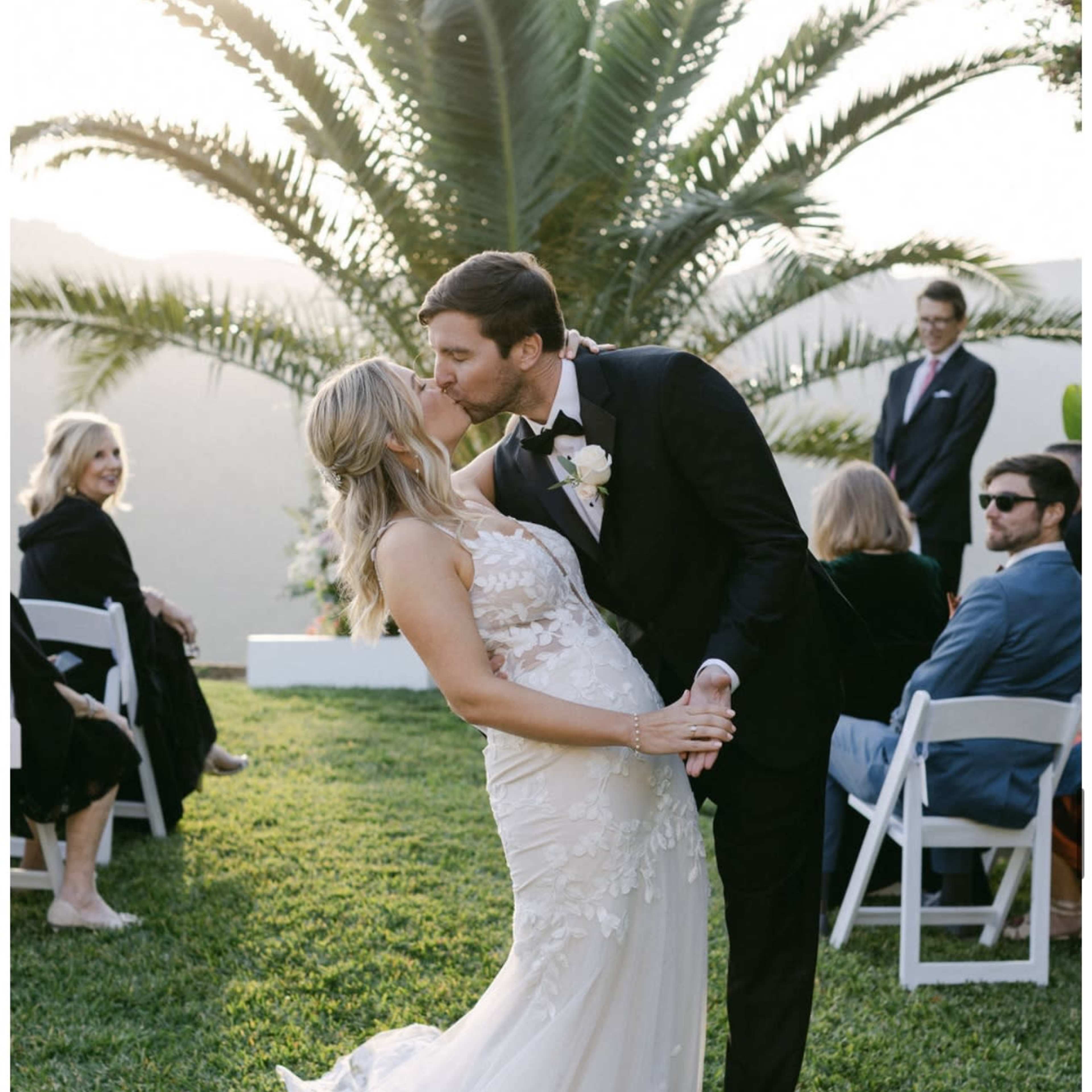 A couple kisses during their outdoor wedding ceremony, surrounded by guests seated on white chairs and a palm tree in the background.