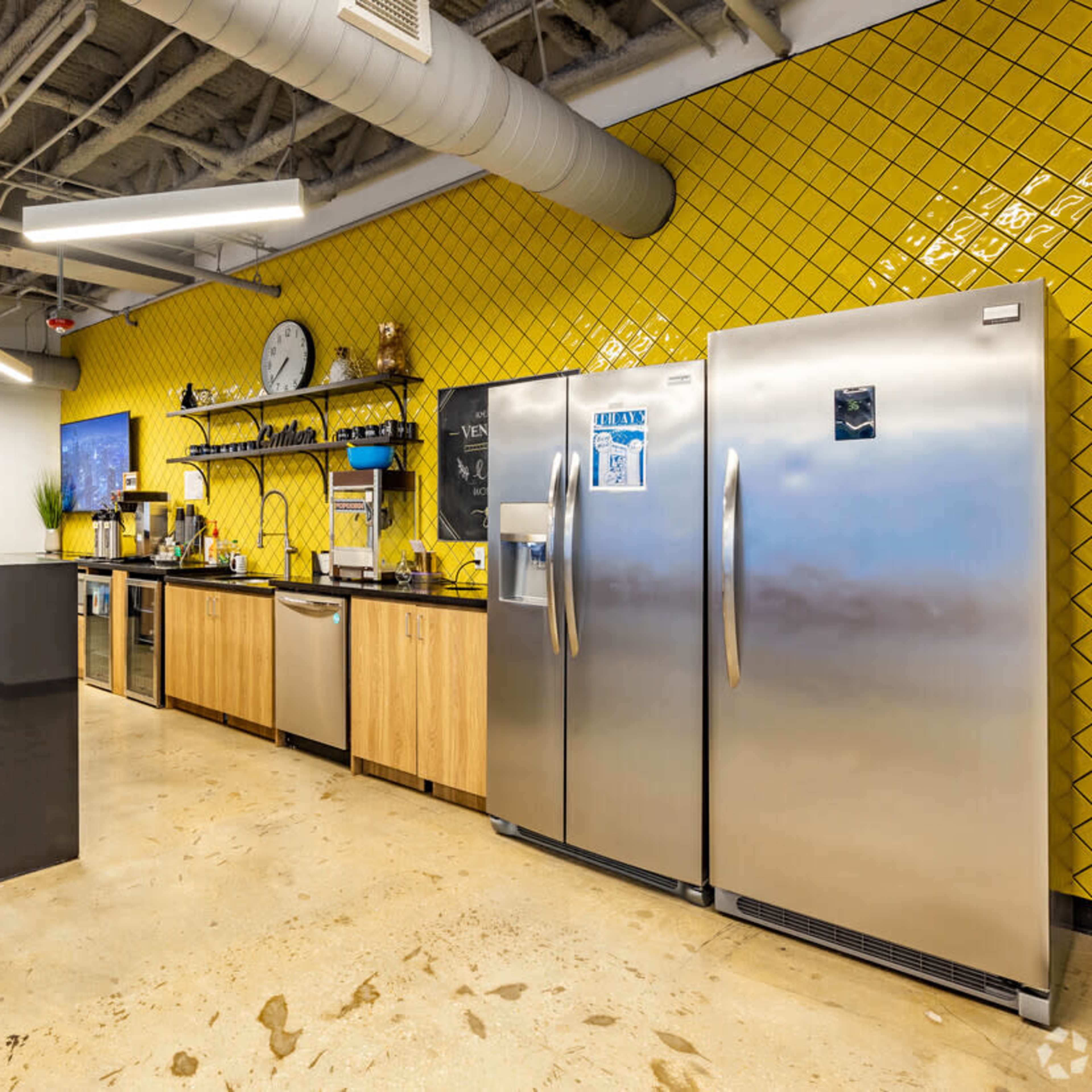 A modern kitchen area featuring stainless steel refrigerators, wooden cabinets, and a black countertop with bar stools, all set against a bright yellow tiled wall.