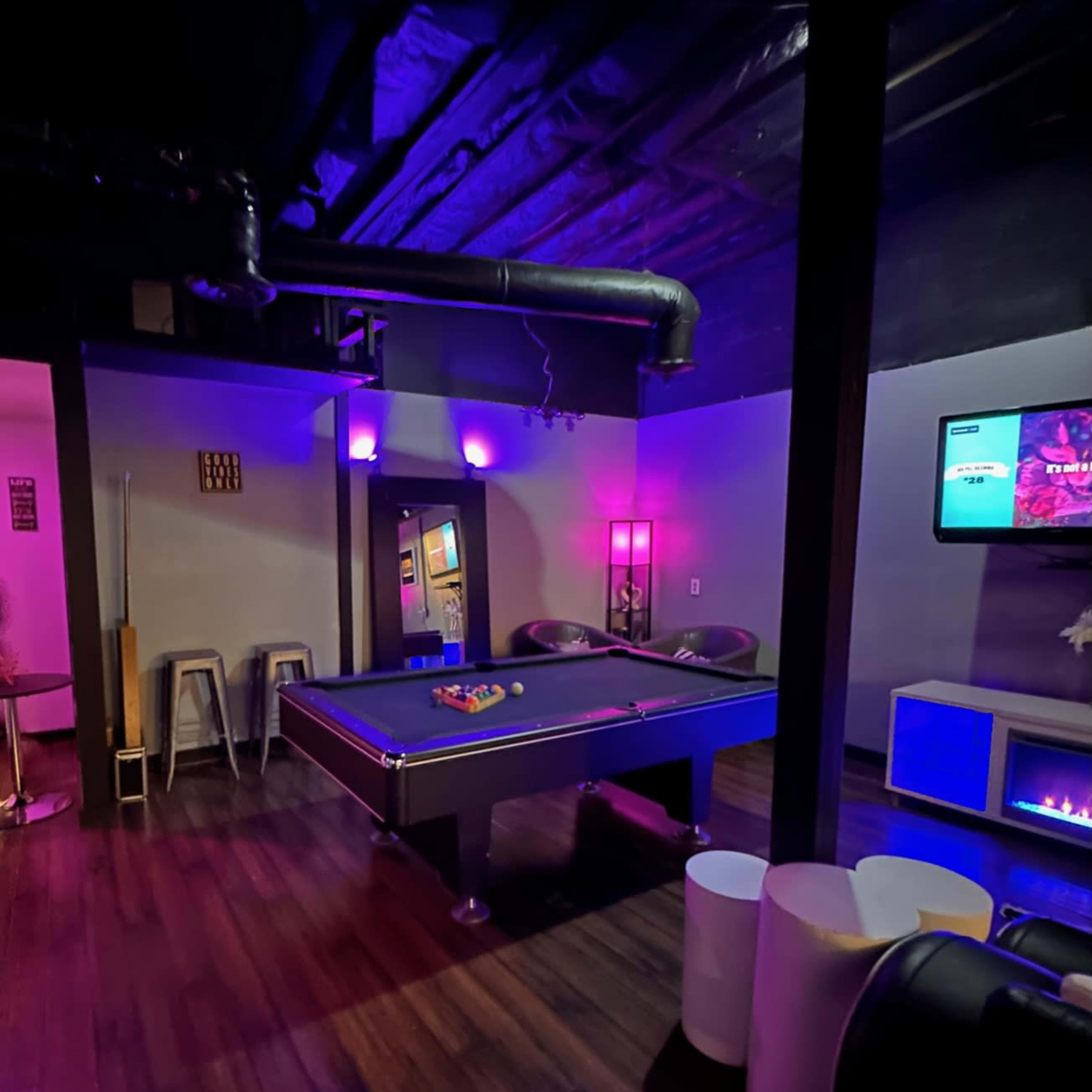 A recreation room featuring a pool table, bar stools, and a television, illuminated with purple lighting.