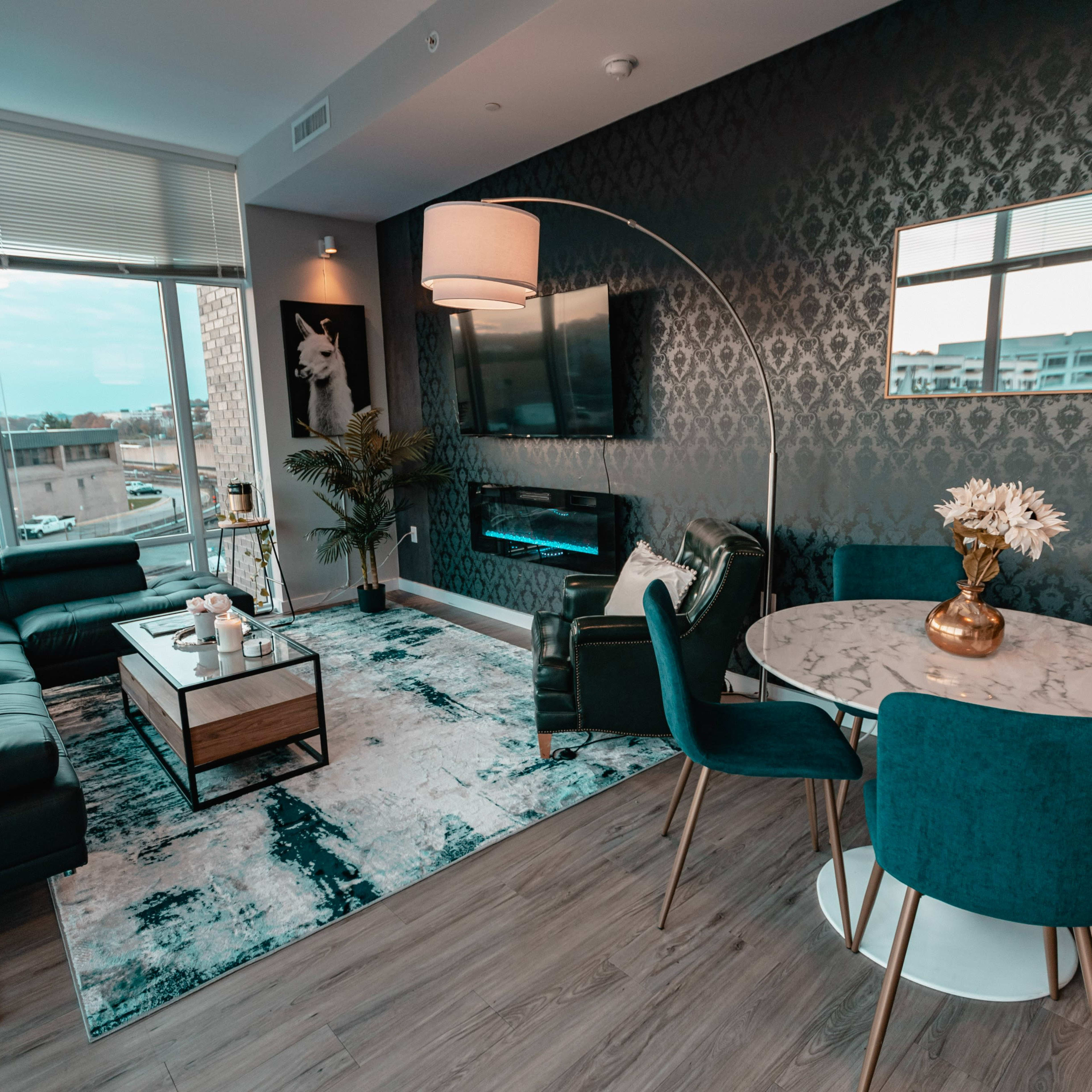 A modern living room featuring a green couch, a round dining table with teal chairs, a marble coffee table, a wall-mounted TV, and large windows that provide natural light.