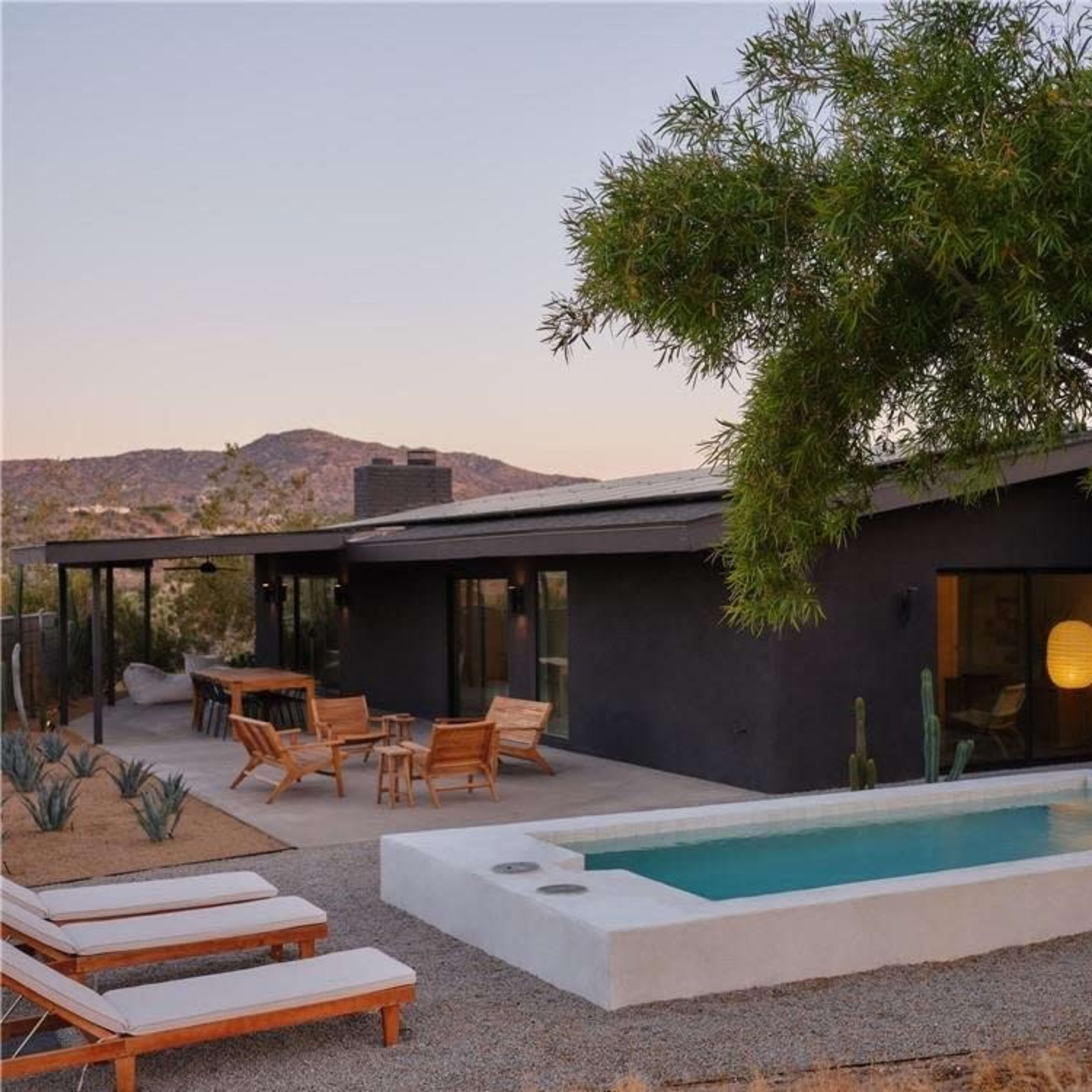 A modern black house with a small pool is surrounded by desert landscaping and mountains in the background.