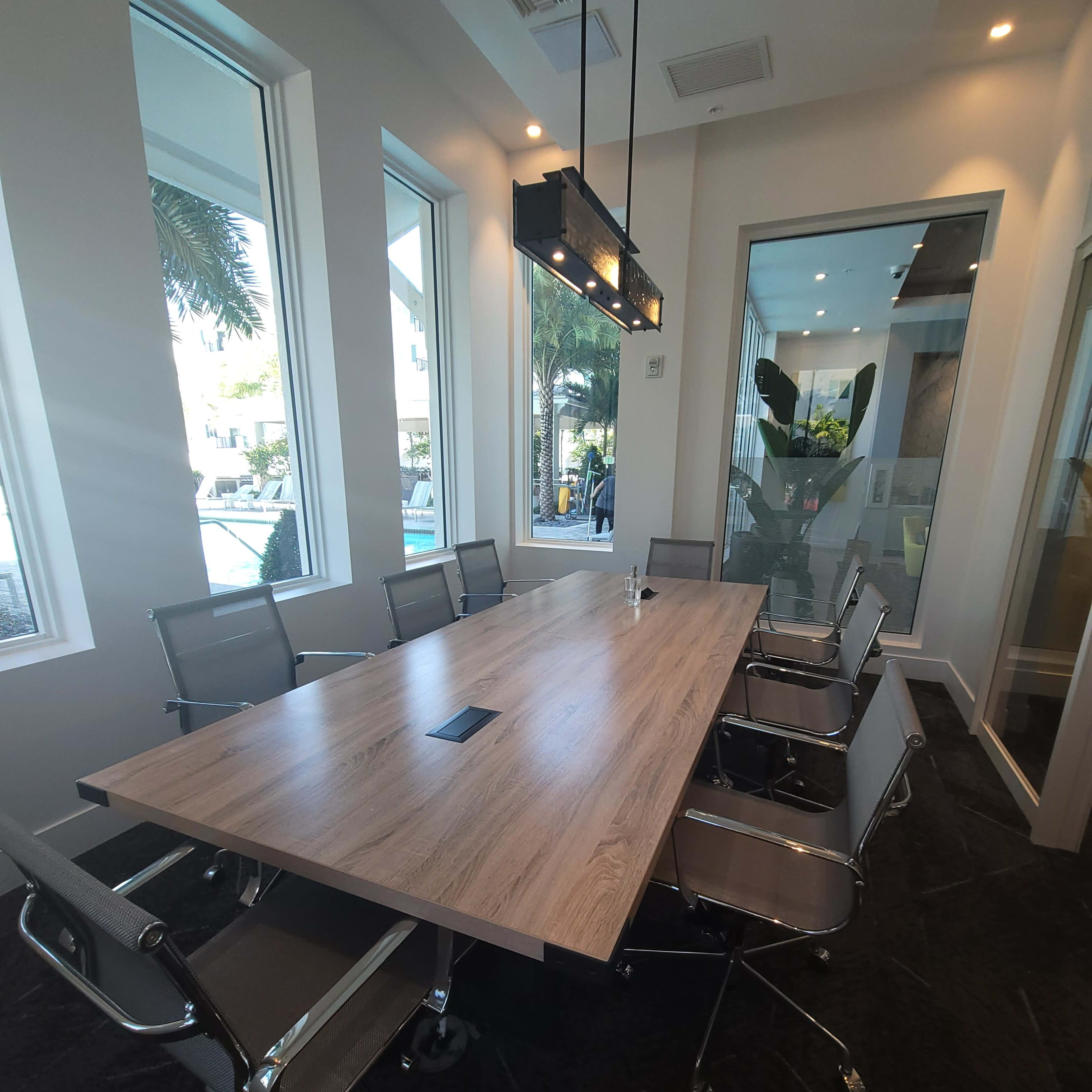 A modern conference room features a large wooden table surrounded by metal chairs, with windows providing natural light and views of greenery outside.