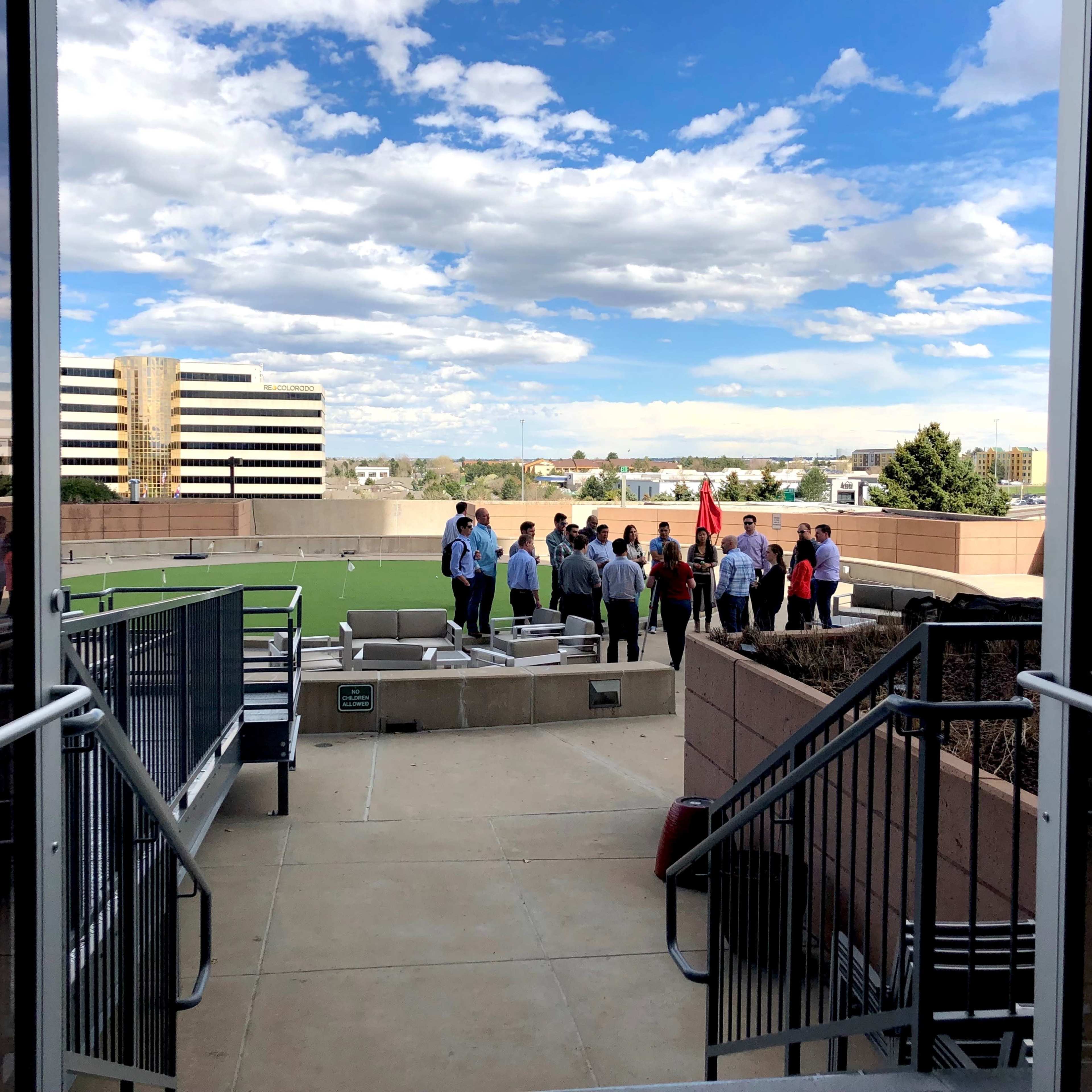A group of people is gathered on a rooftop terrace with a green area and buildings in the background under a cloudy sky.