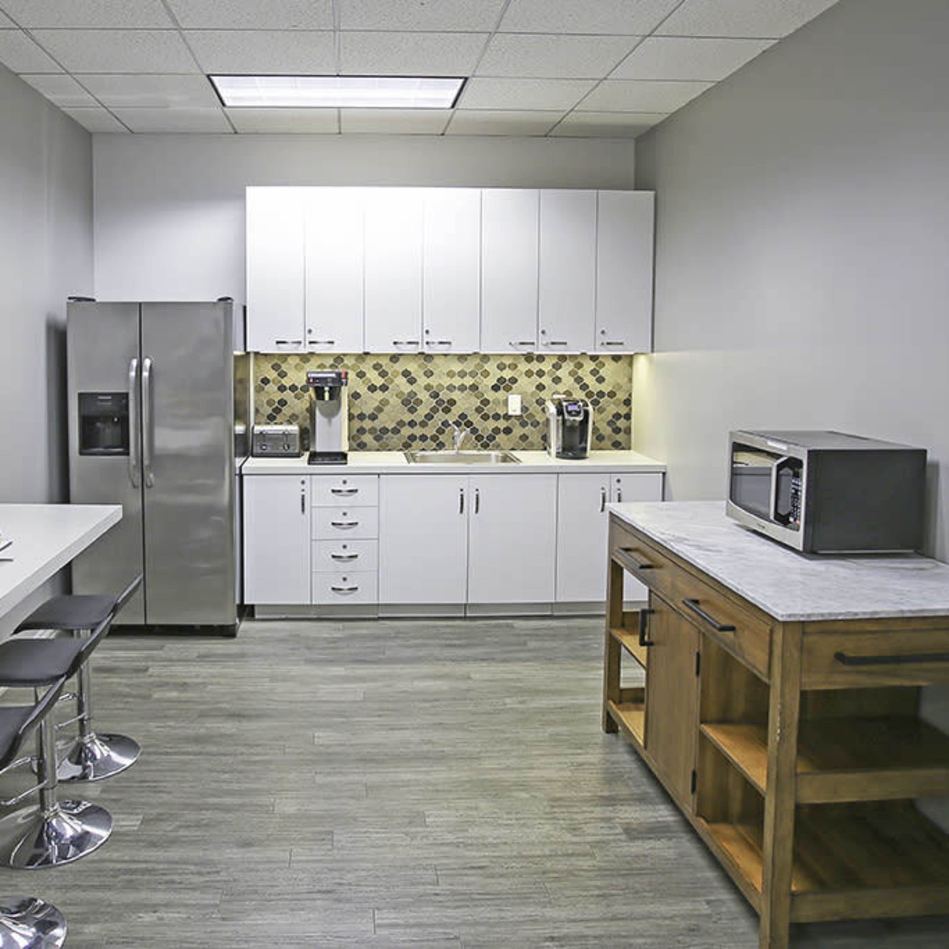 A modern kitchenette features a stainless steel refrigerator, white cabinets, a countertop with a coffee maker, and a snack bar with stools.