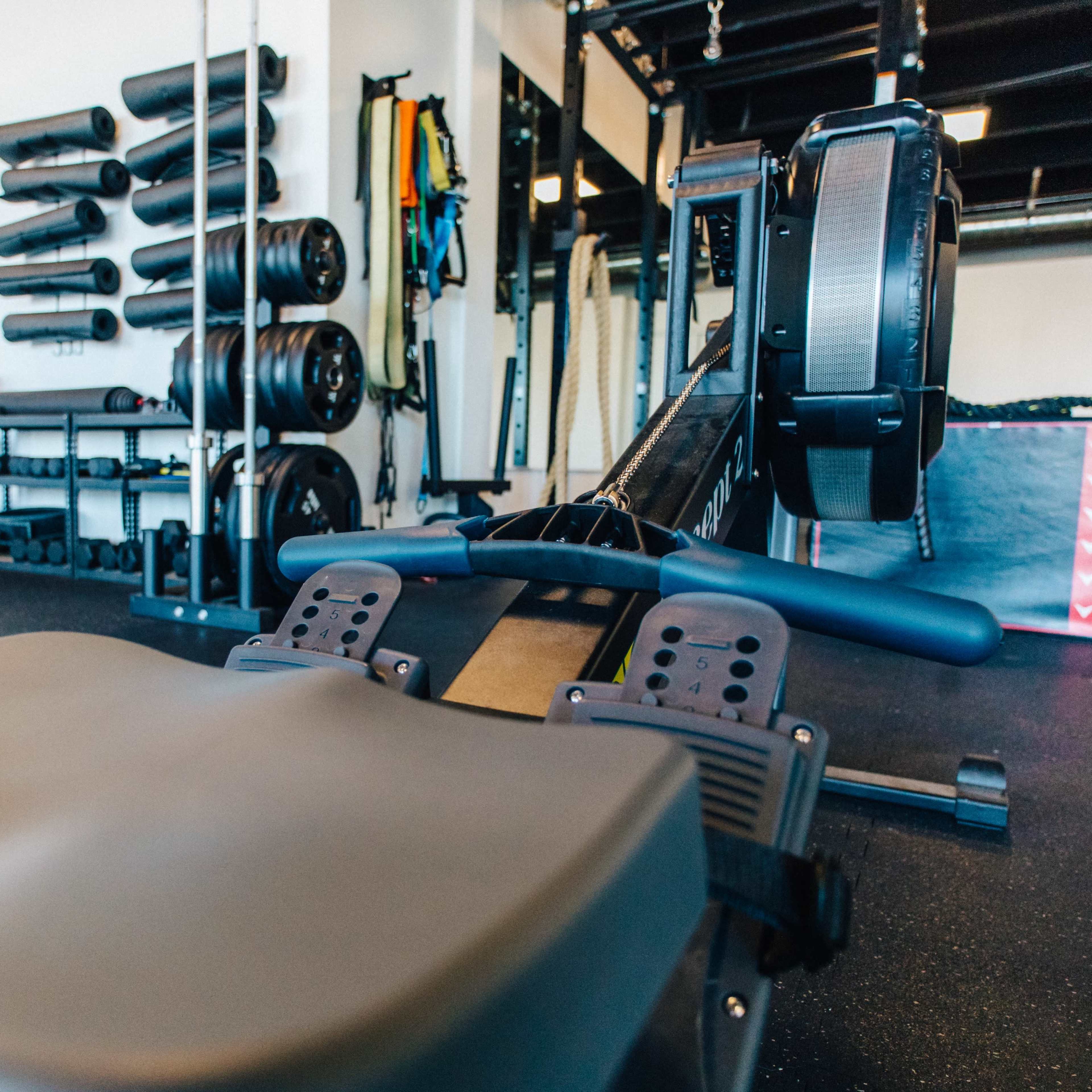 A rowing machine in a gym, with free weights and exercise equipment arranged on the walls in the background.