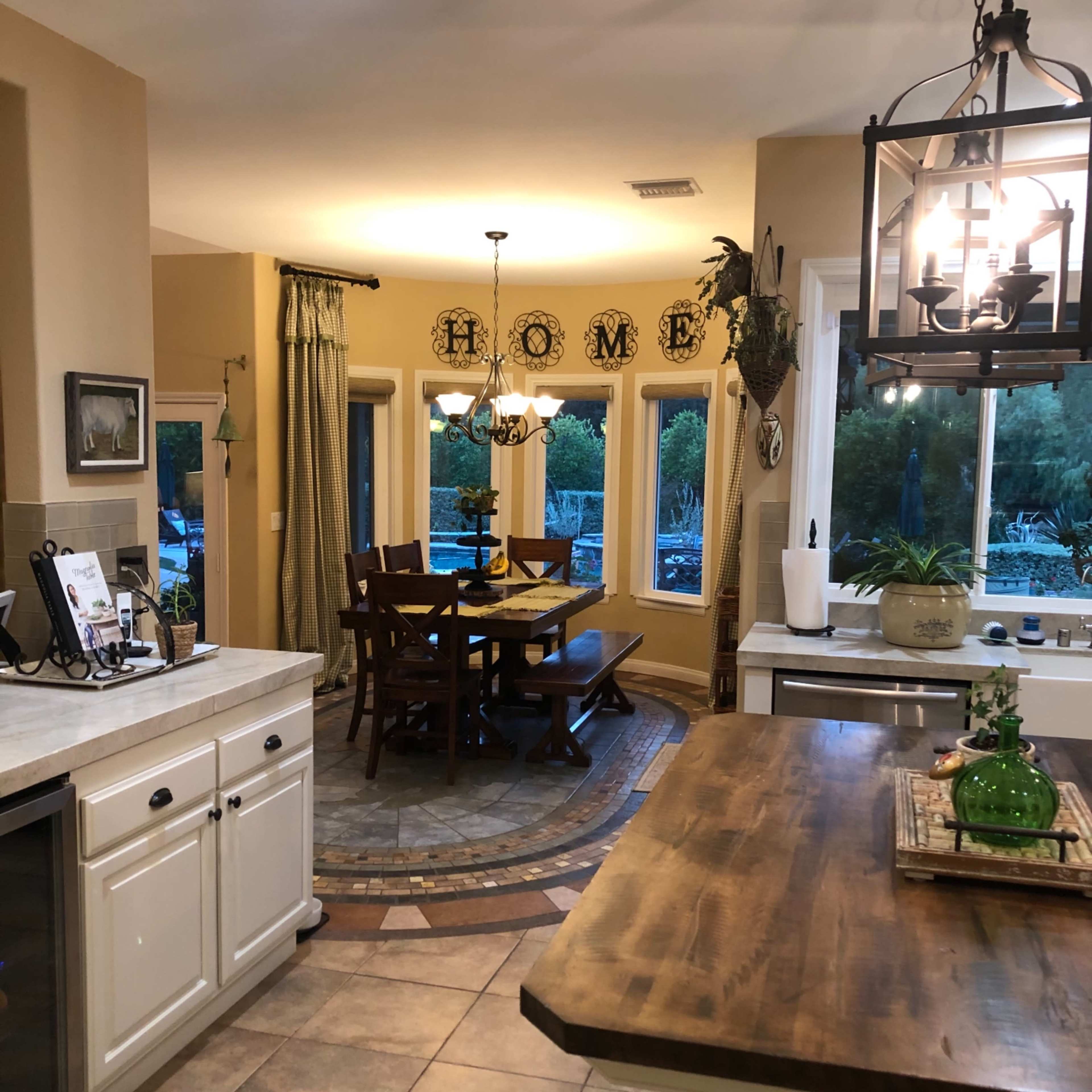 A well-lit kitchen features a dining area with a wooden table surrounded by chairs and large windows, showcasing a homey atmosphere.