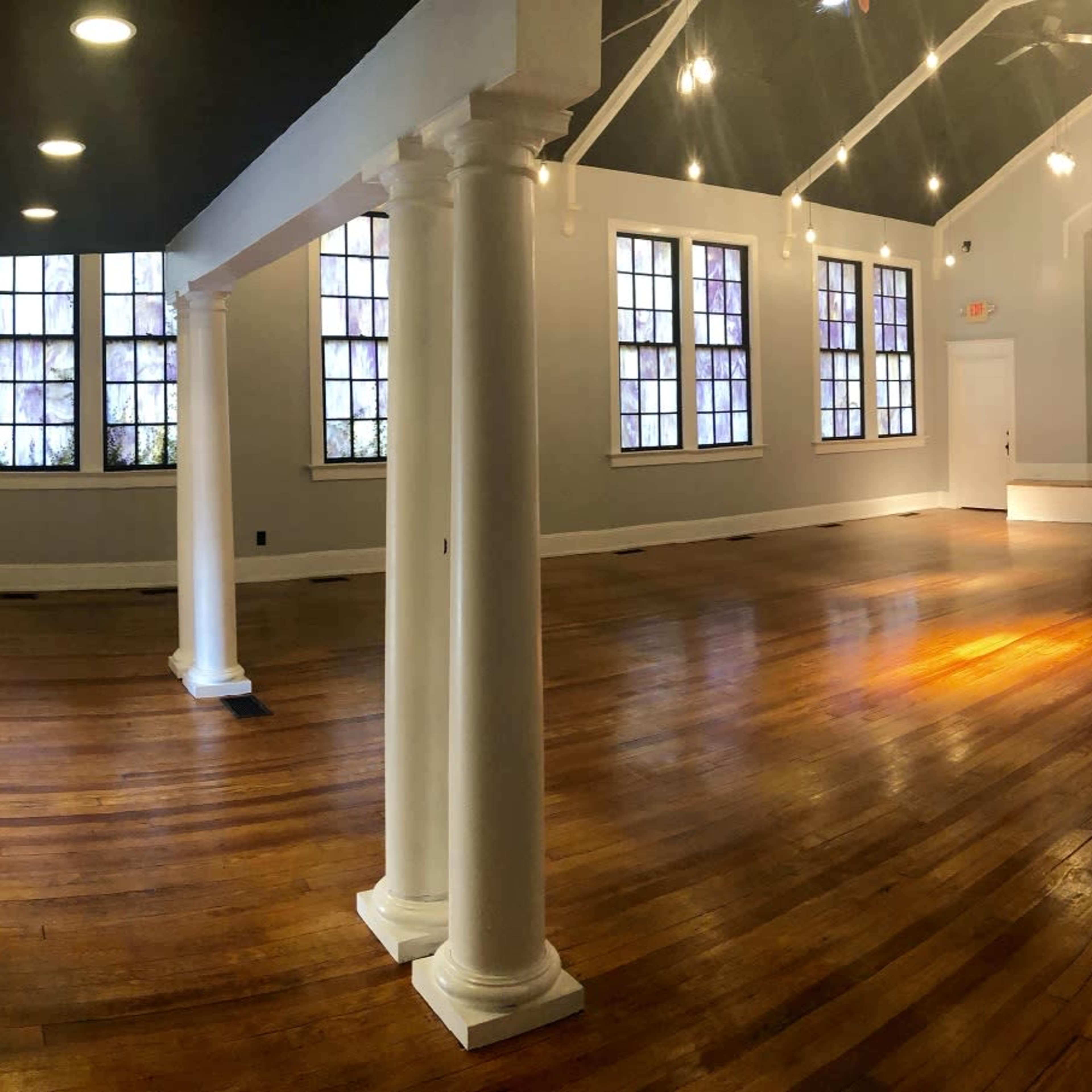 A spacious interior room with wooden floors, white walls, and columns, featuring large windows and a raised stage area at the far end.