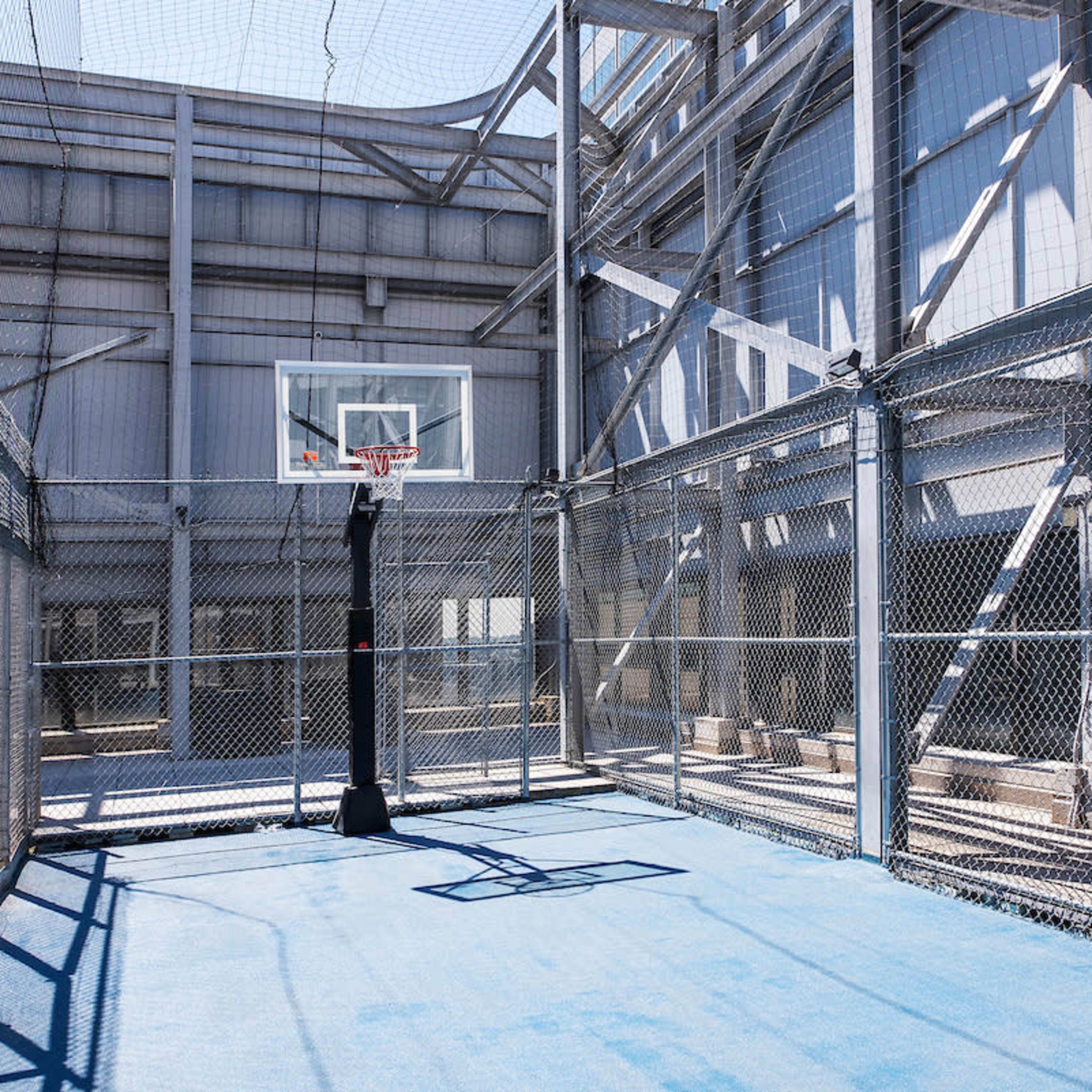 A fenced basketball court with a hoop and a blue playing surface, situated within a metal structure.