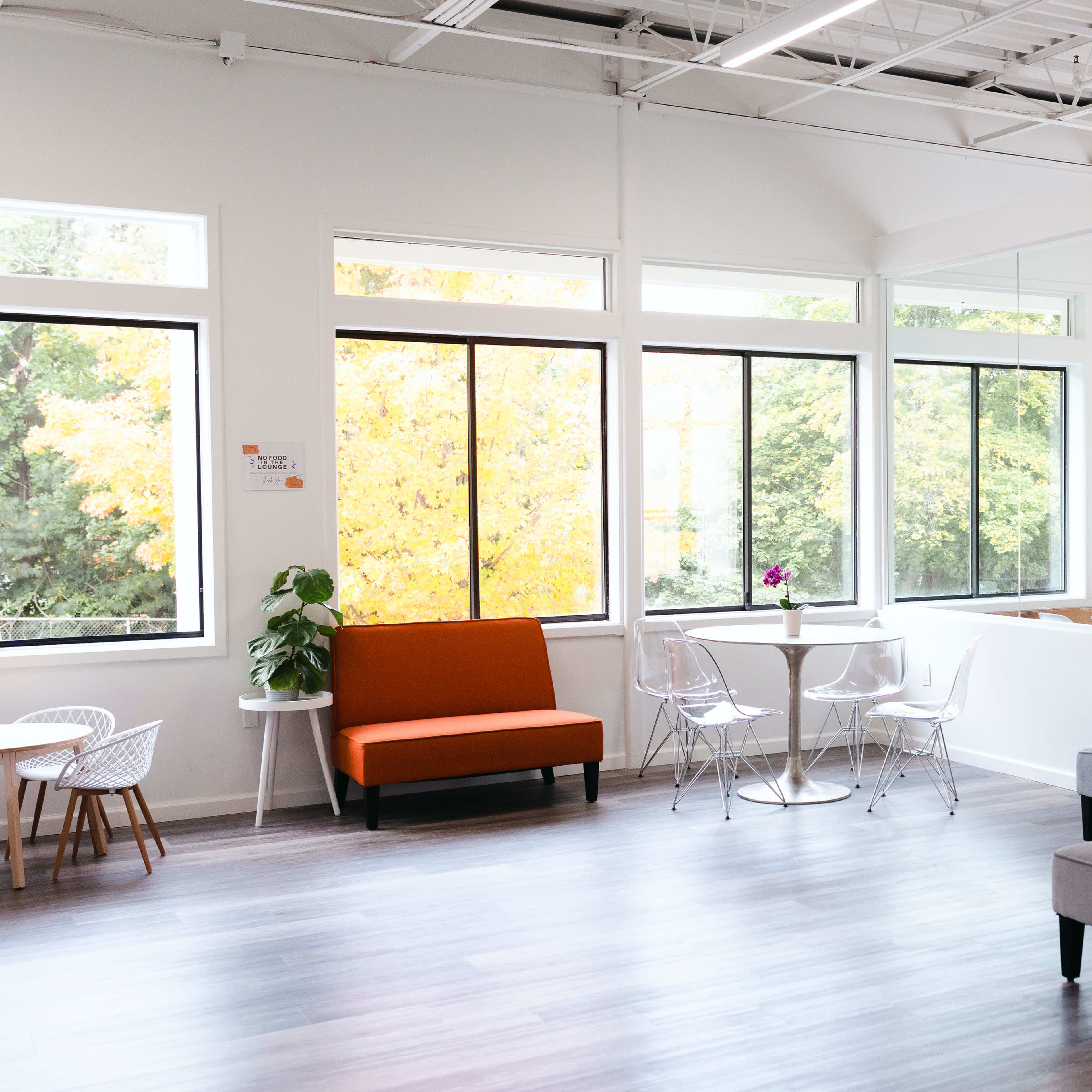 A bright, modern office space with large windows, a gray sofa, an orange chair, and a small table with clear chairs.