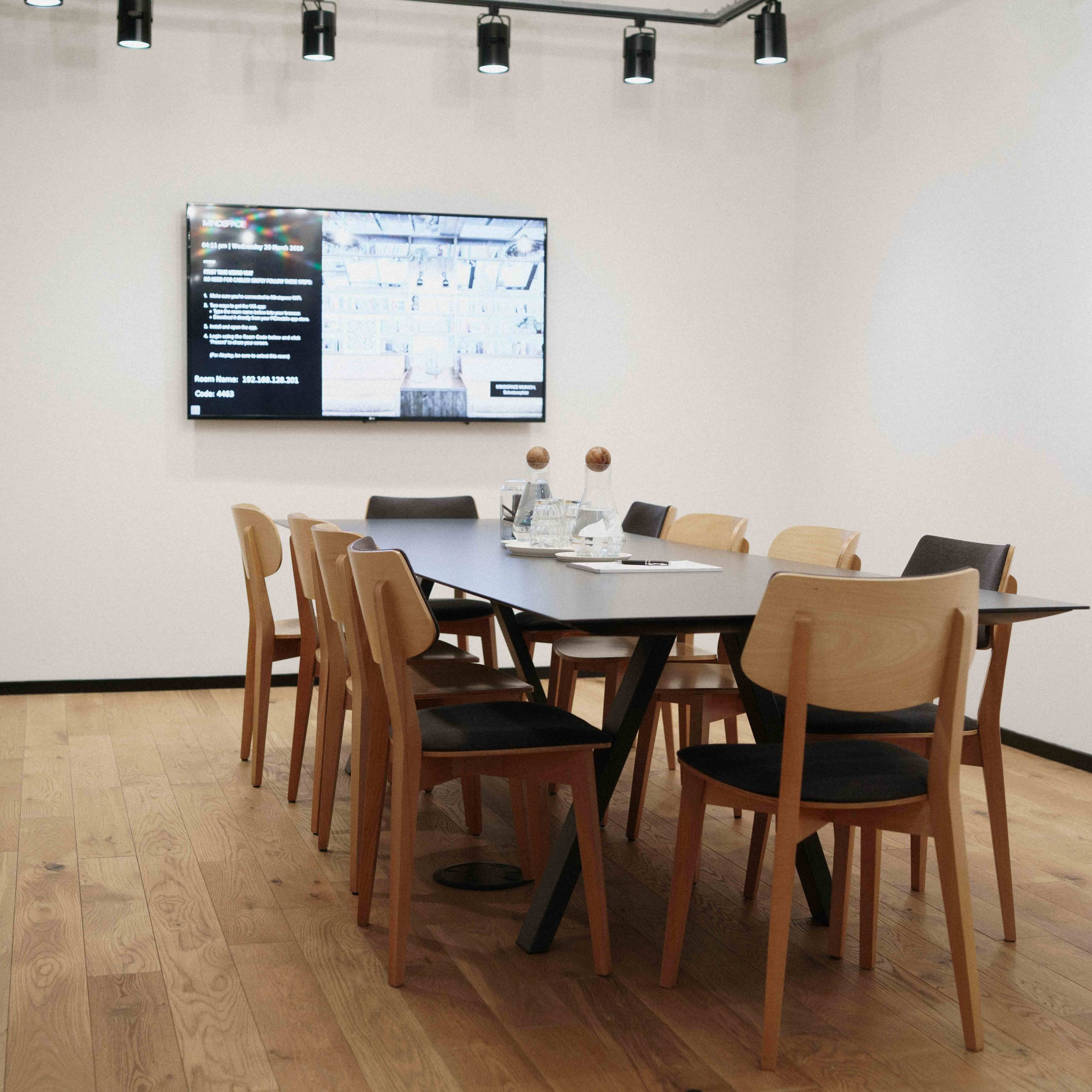 A modern meeting room features a long table surrounded by wooden chairs and a large screen displaying information on a white wall.