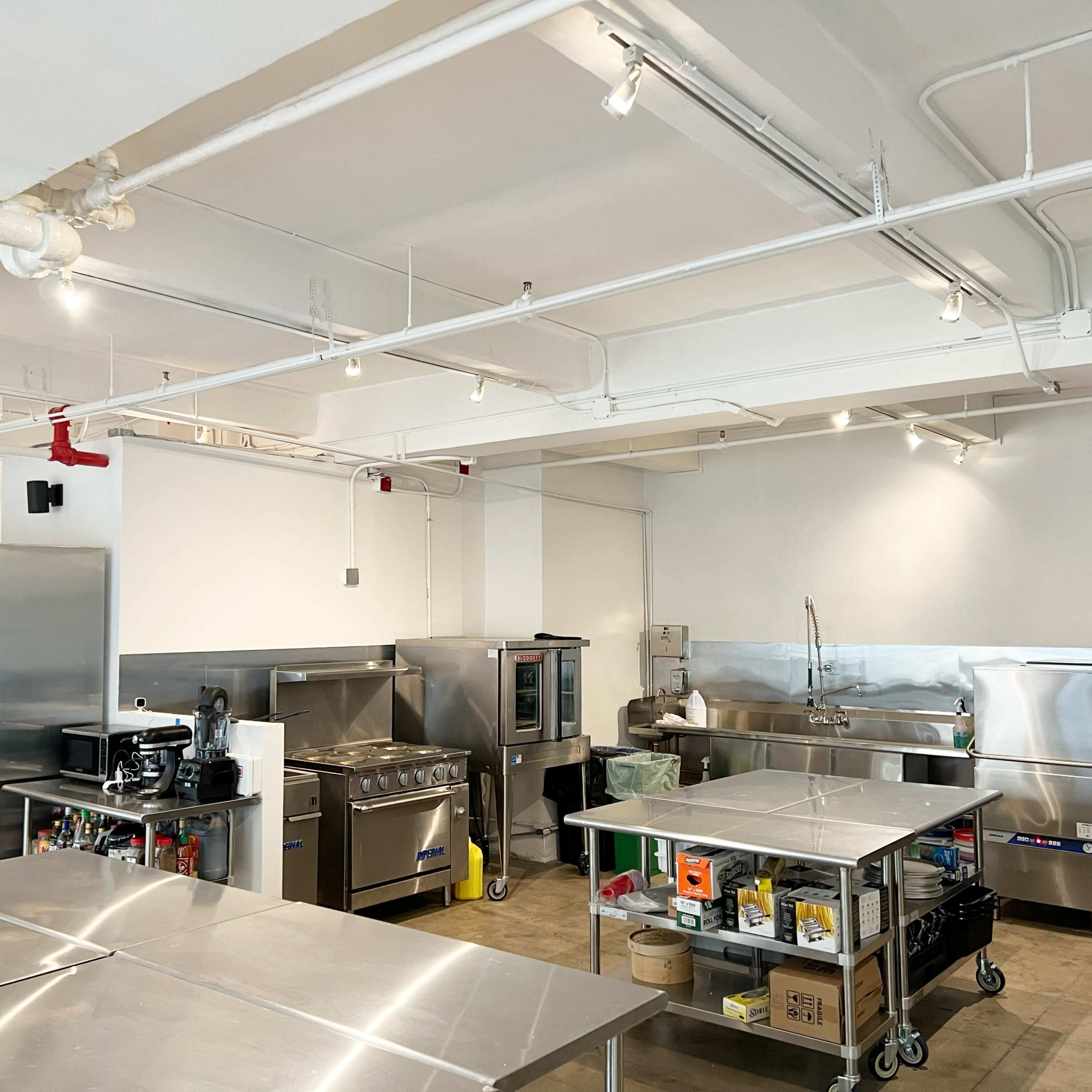 A commercial kitchen features stainless steel appliances, stainless steel tables, and a spacious layout with overhead lighting.