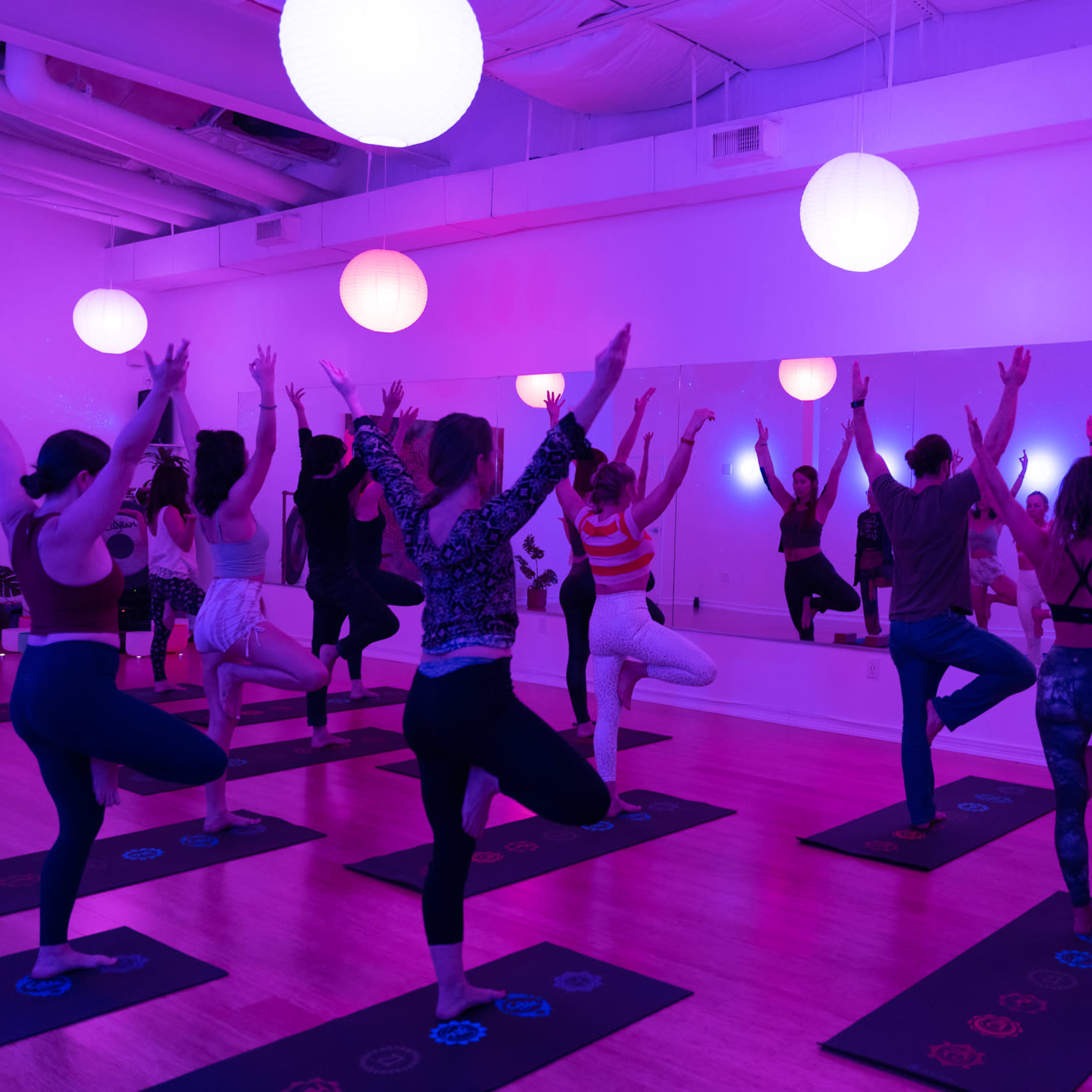 A group of people practices yoga in a brightly lit studio with purple and white hanging lamps.
