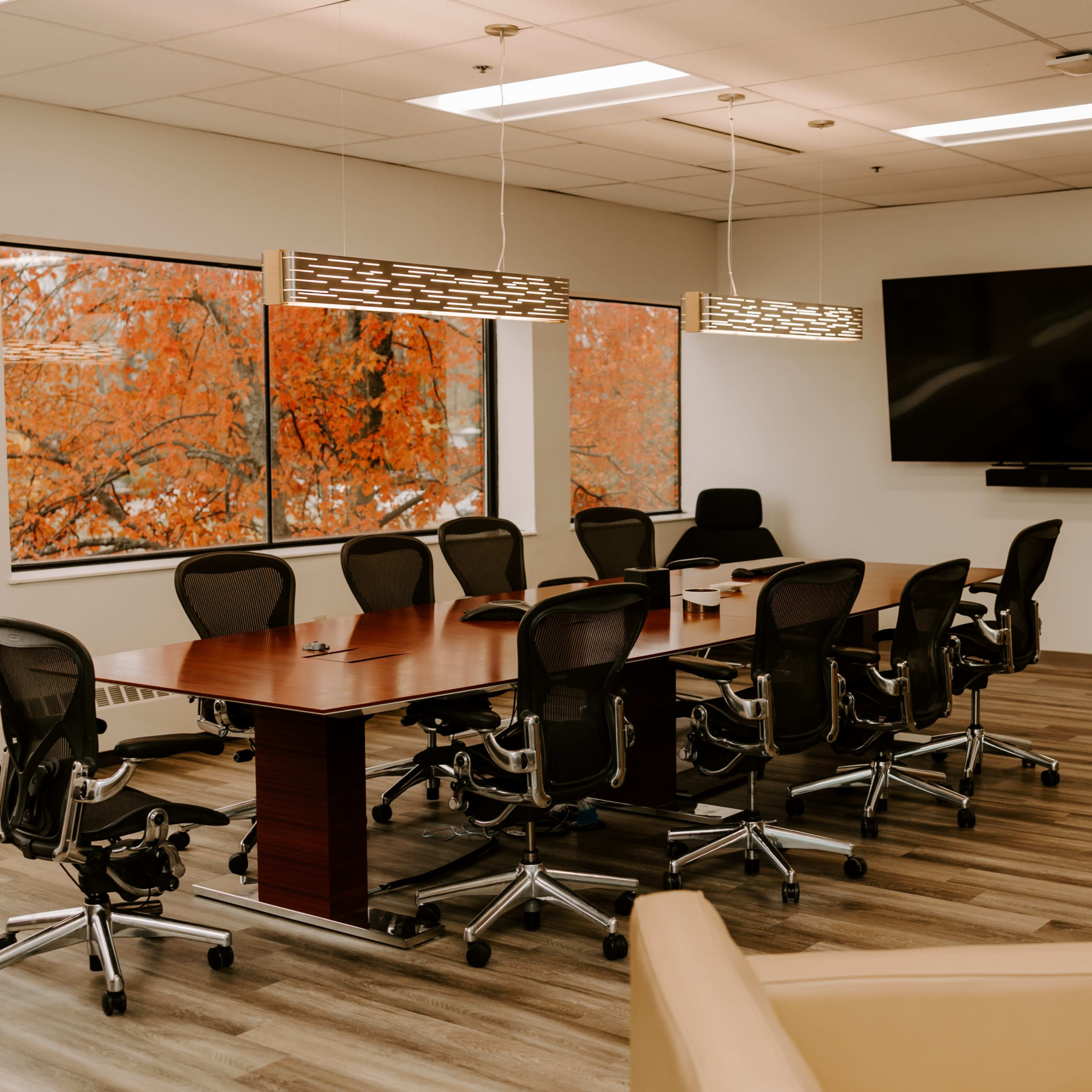 A conference room features a large wooden table surrounded by ergonomic chairs, with large windows displaying fall foliage outside.