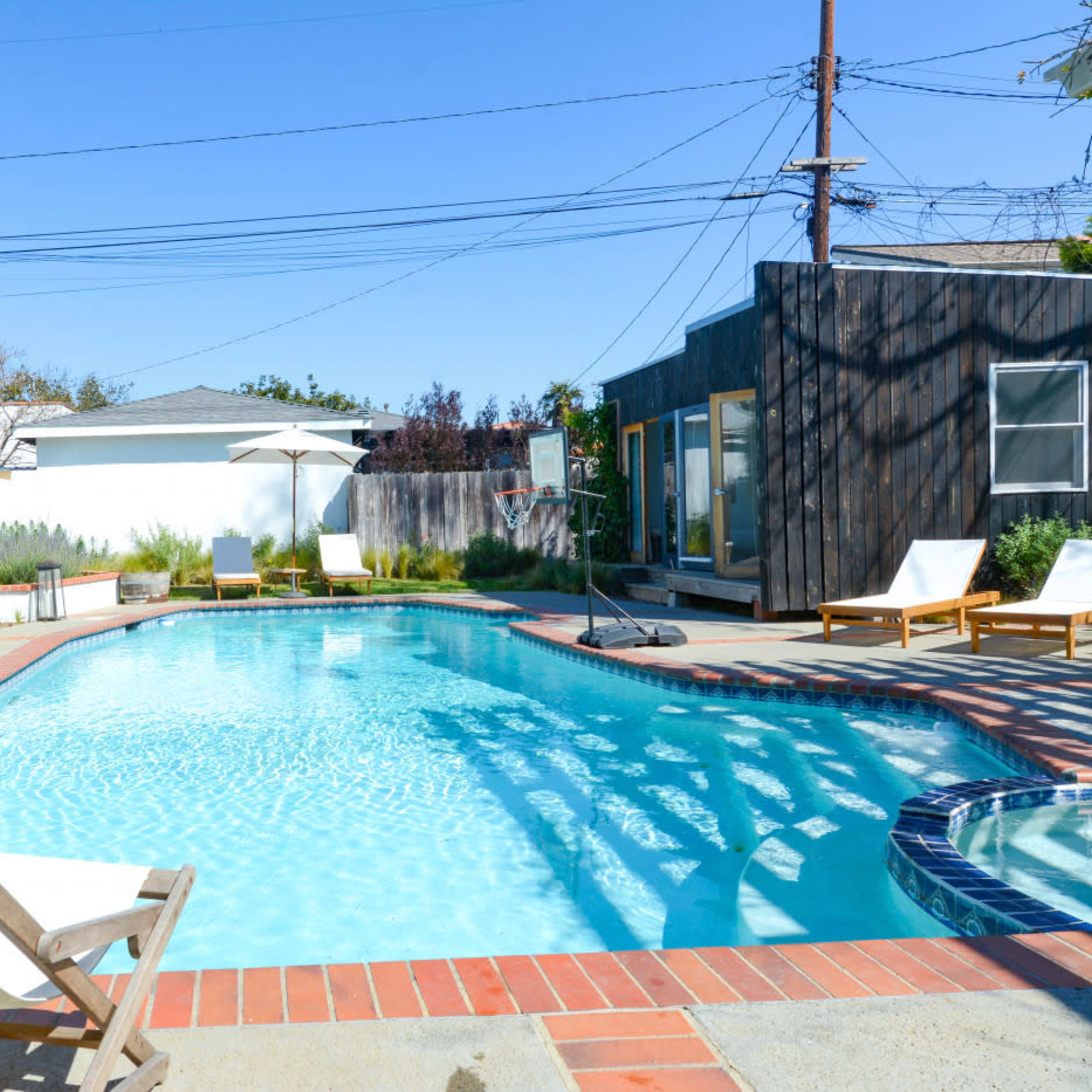A blue swimming pool with lounge chairs nearby, surrounded by a landscaped area and a wooden structure in the background.