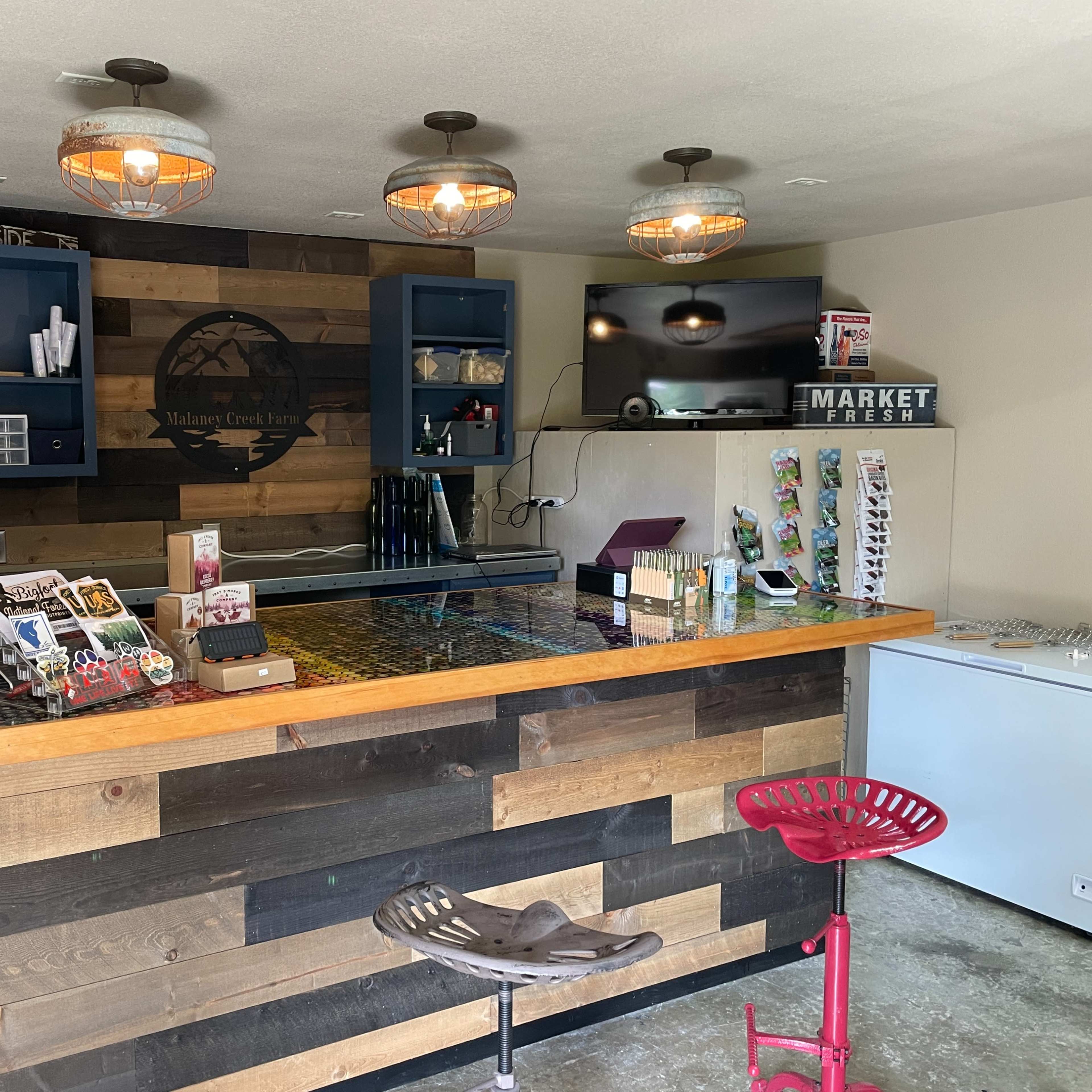 A small, rustic bar area with a wooden counter, shelves displaying various items, and a television mounted on the wall.