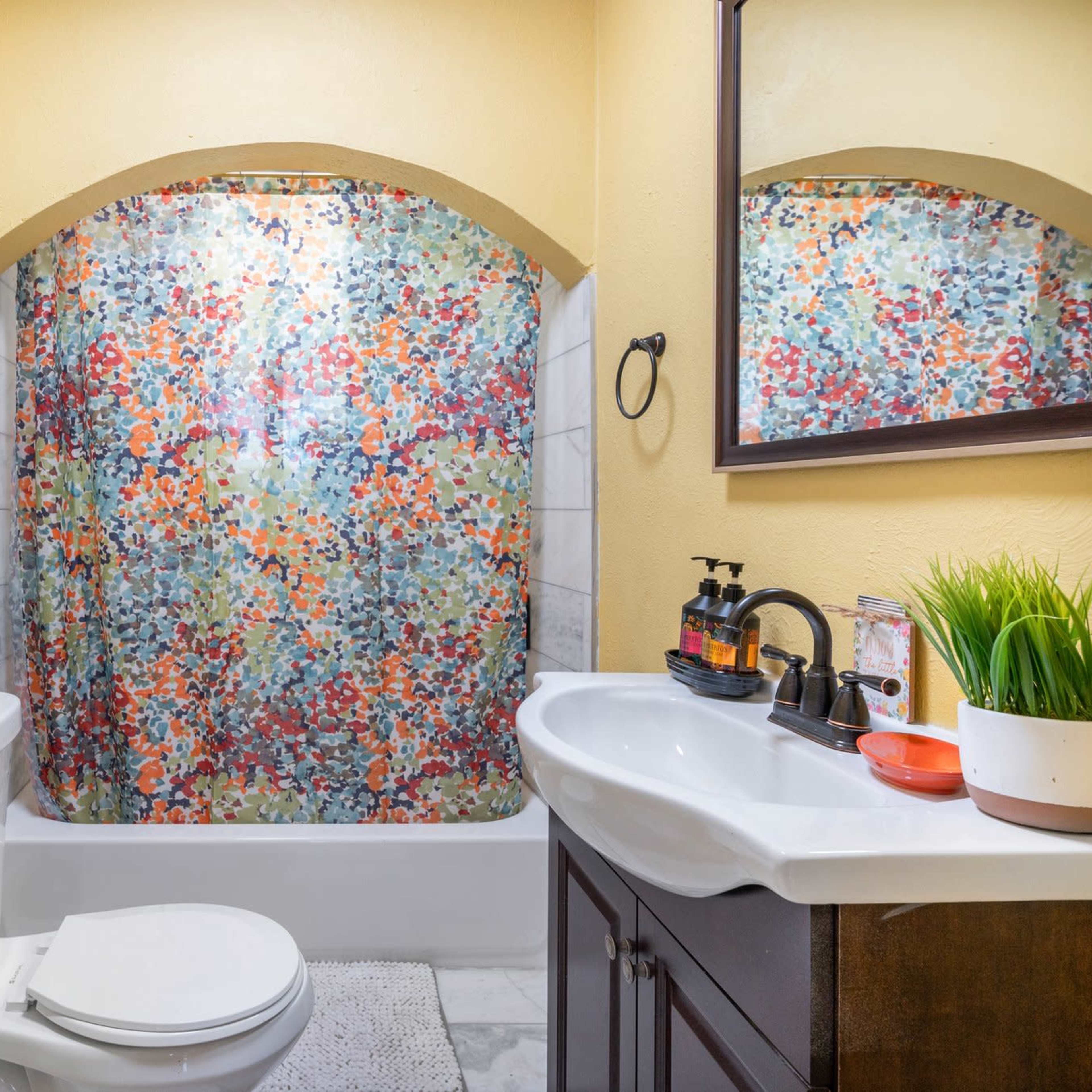 The bathroom features a colorful floral shower curtain, a white toilet, and a sink with a mirror and decorative items.