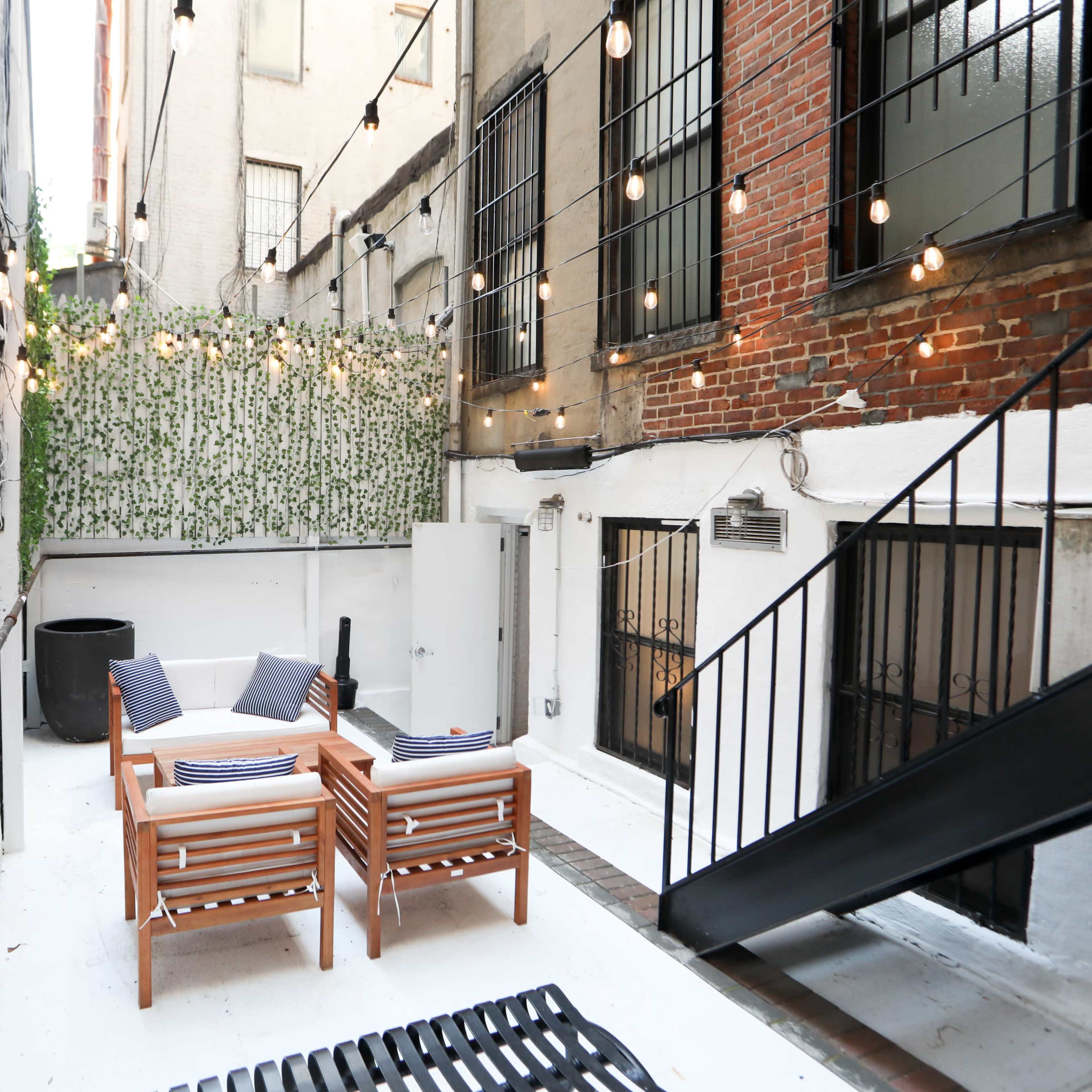 A small outdoor patio area featuring two wooden chairs with cushions, surrounded by white walls, string lights, and a staircase leading to a higher level.