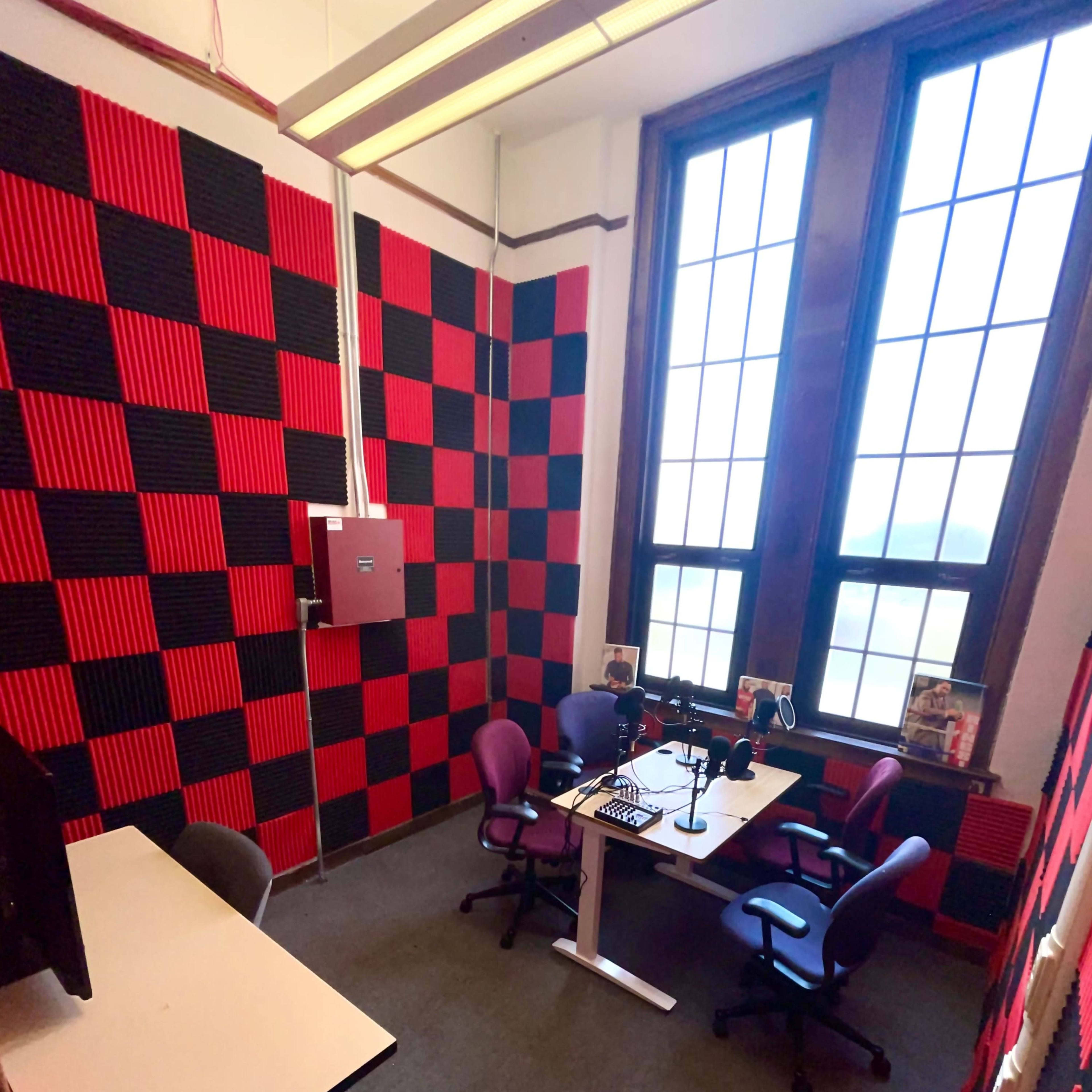 A small, soundproofed room with black and red checkered walls, two chairs, a desk, and equipment for recording or broadcasting.