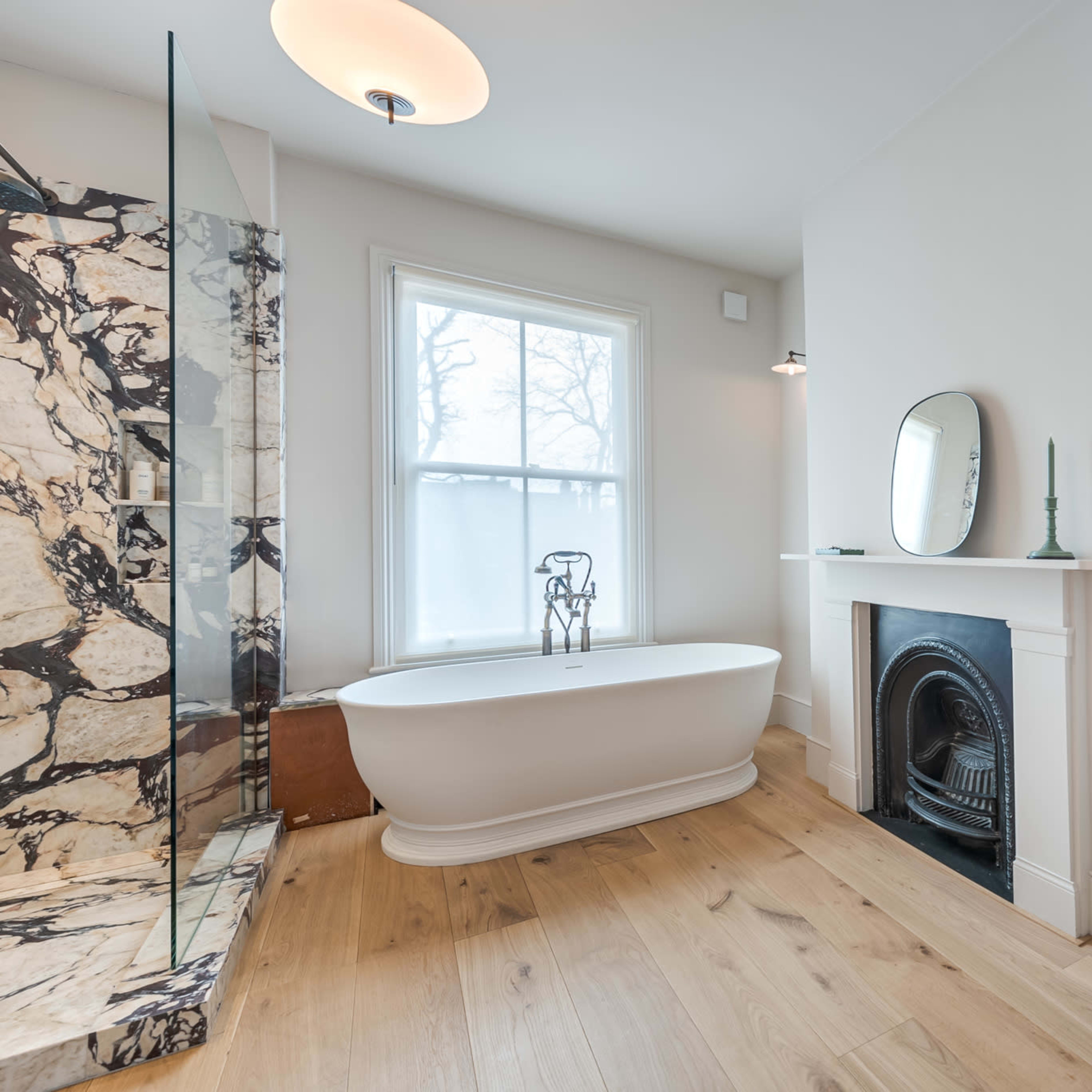 A modern bathroom features a freestanding bathtub, a glass-enclosed shower, and a marble sink with a mirror, all illuminated by natural light from a nearby window.