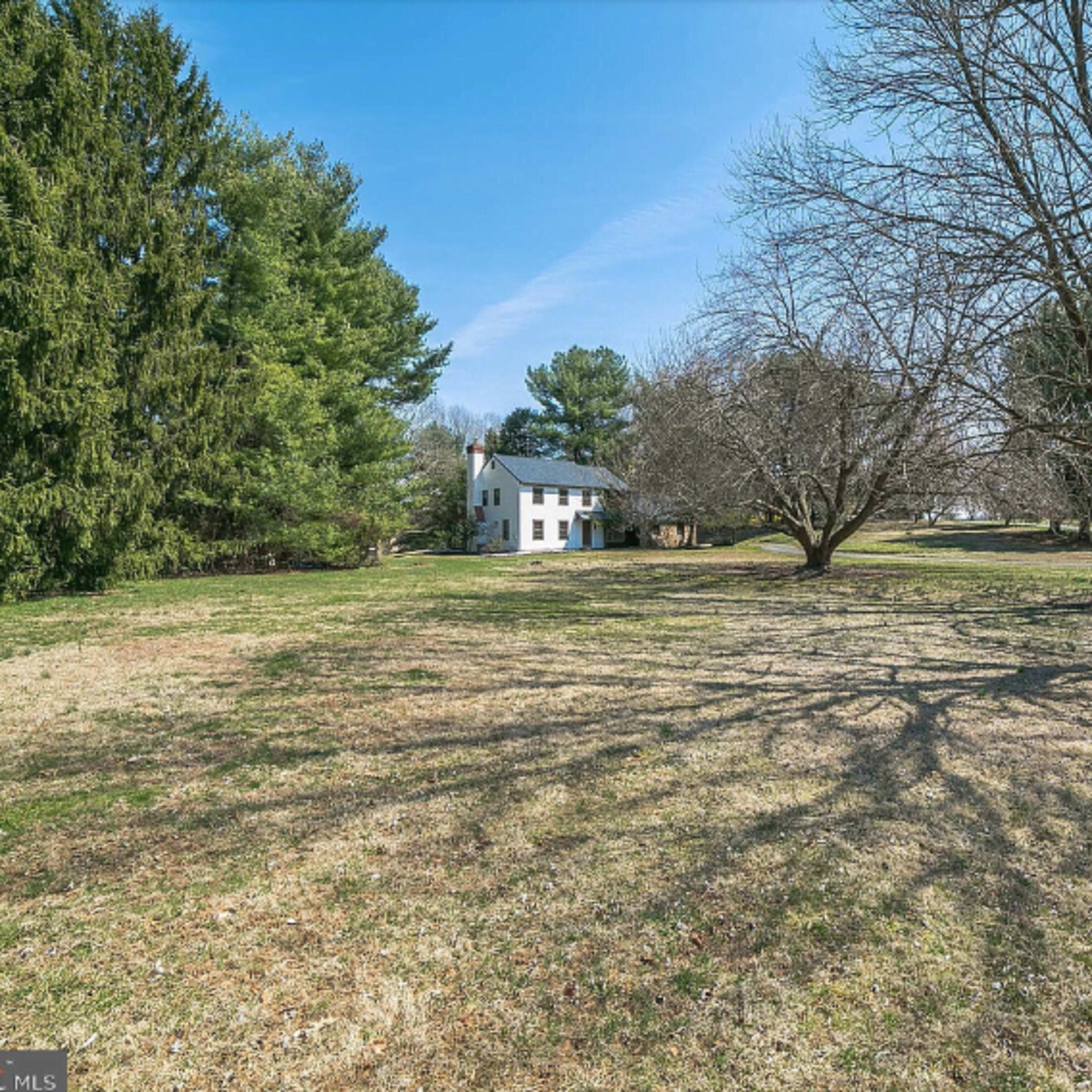 A white house stands in a grassy yard surrounded by several trees under a clear blue sky.