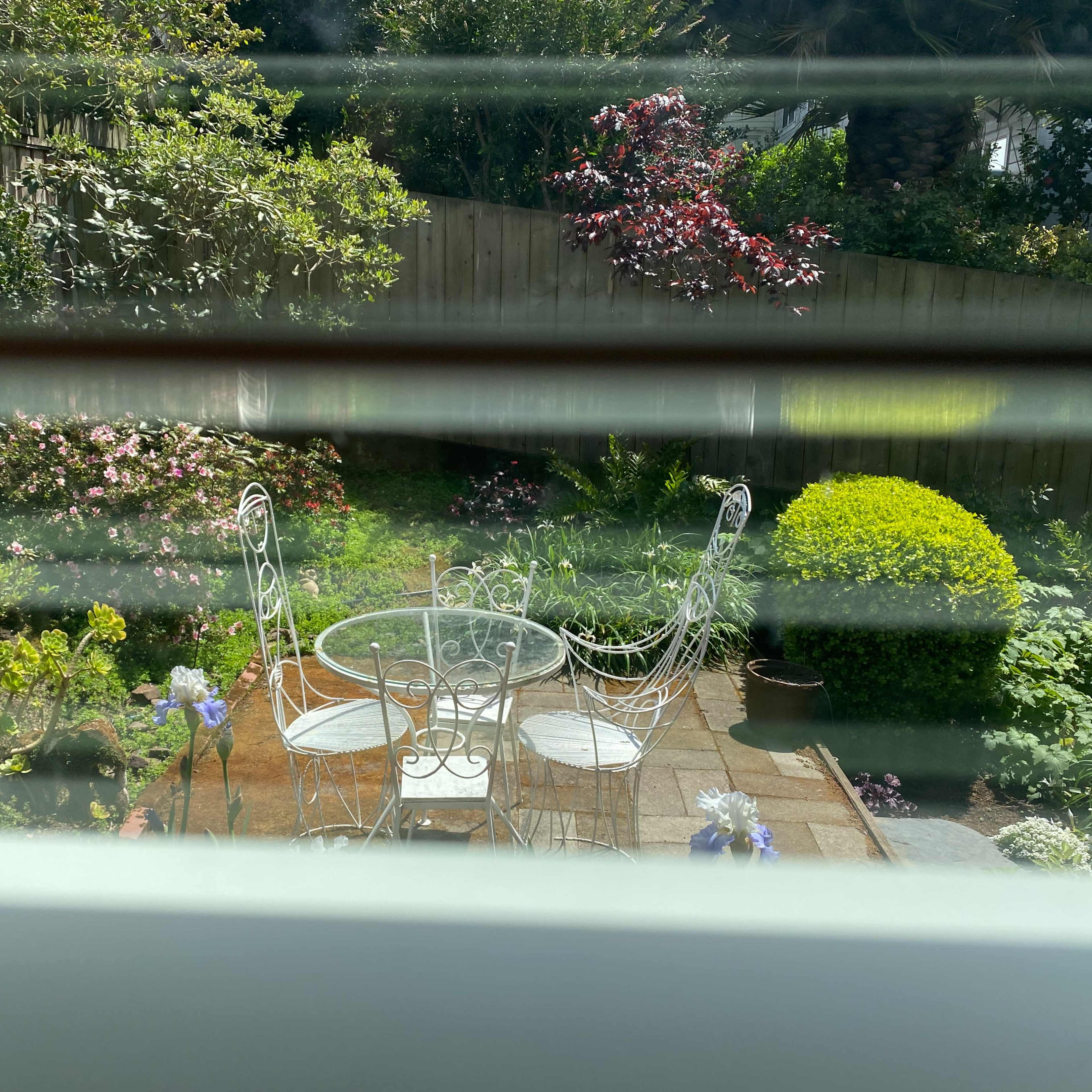 A small outdoor garden features a round table and two chairs surrounded by blooming plants and shrubs.