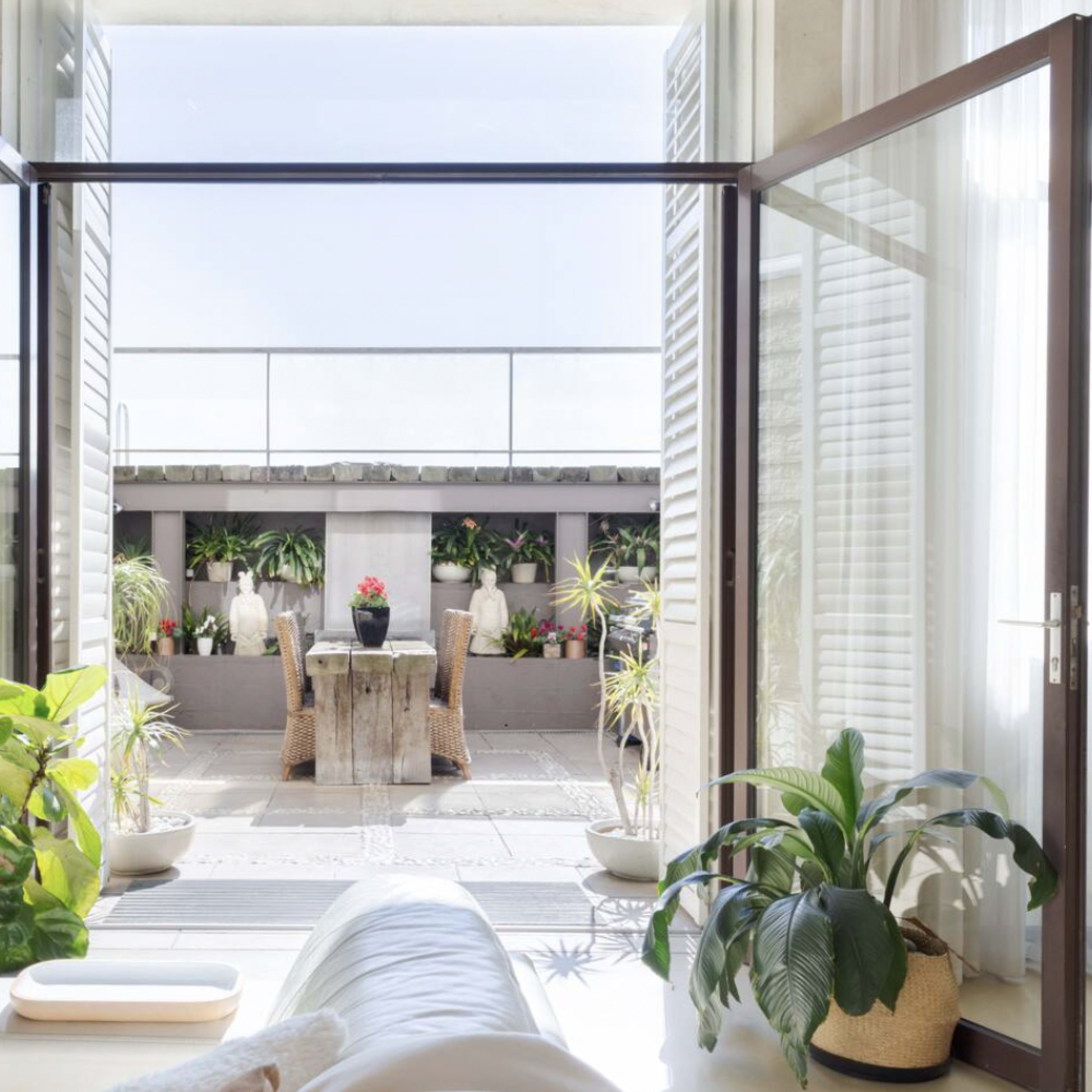 A bright and airy interior space with open glass doors leading to a terrace filled with potted plants.