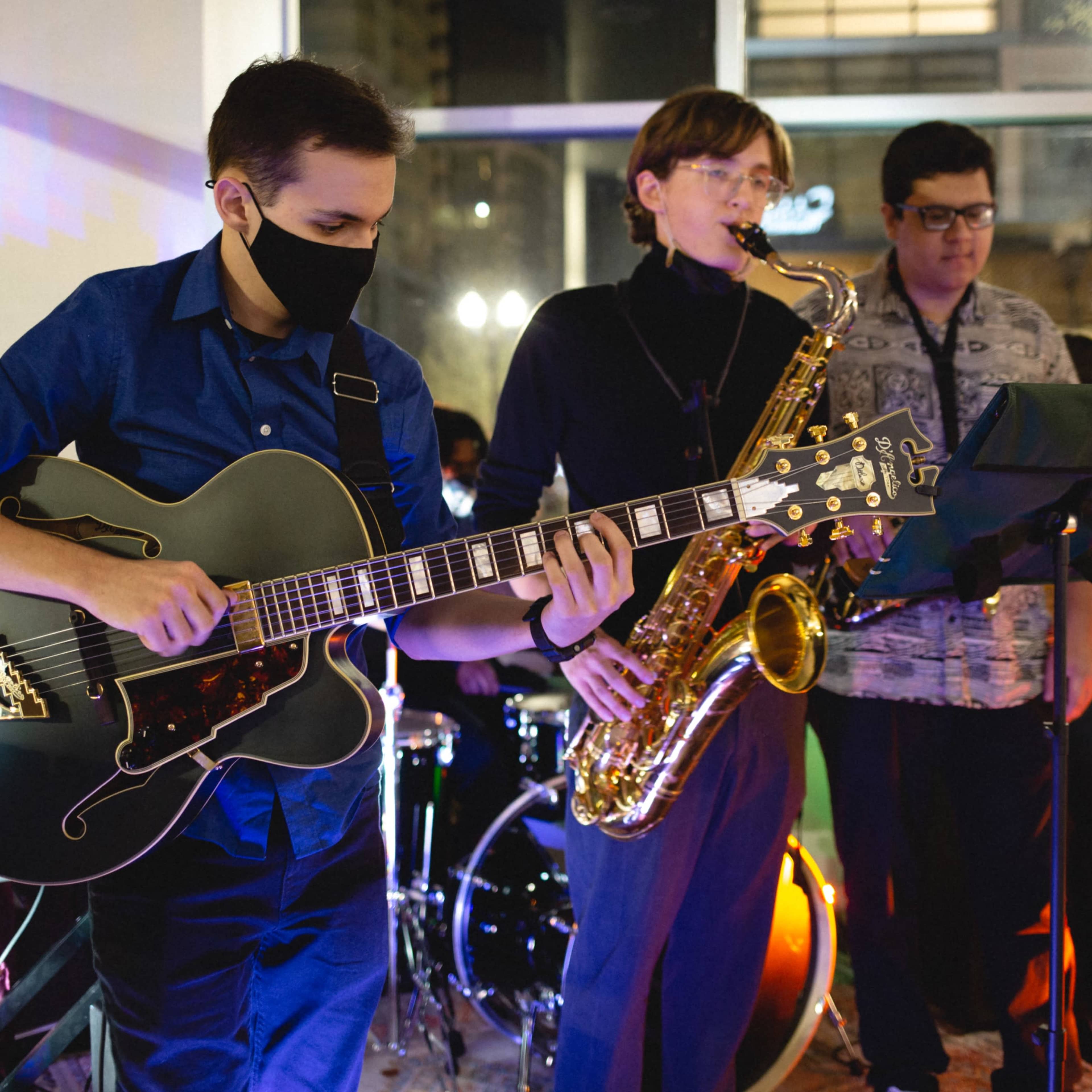A group of musicians performs together in a well-lit venue, featuring a guitarist, a saxophonist, and other band members with various instruments.