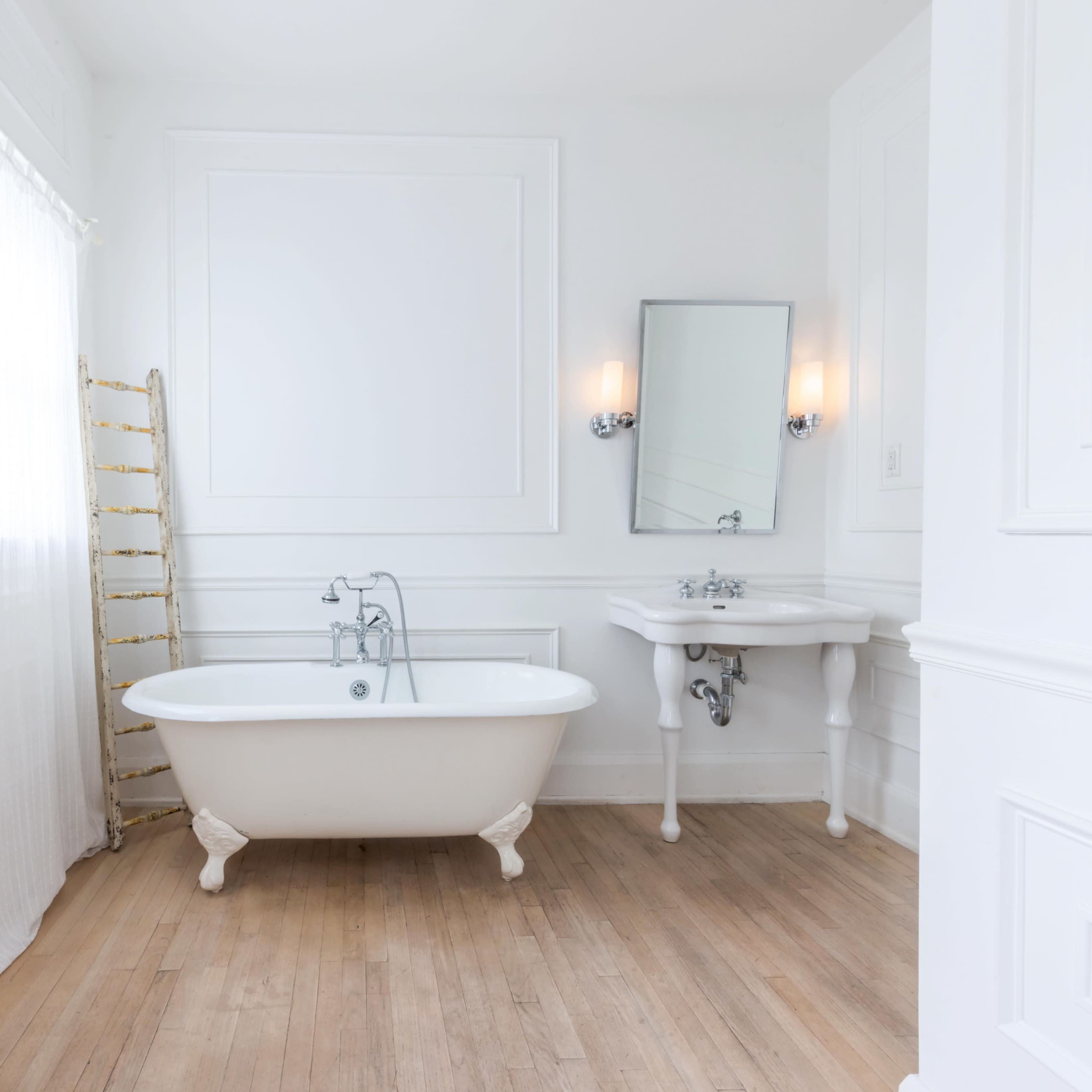 A bright, minimalist bathroom featuring a freestanding bathtub, a small sink, and a wooden floor.