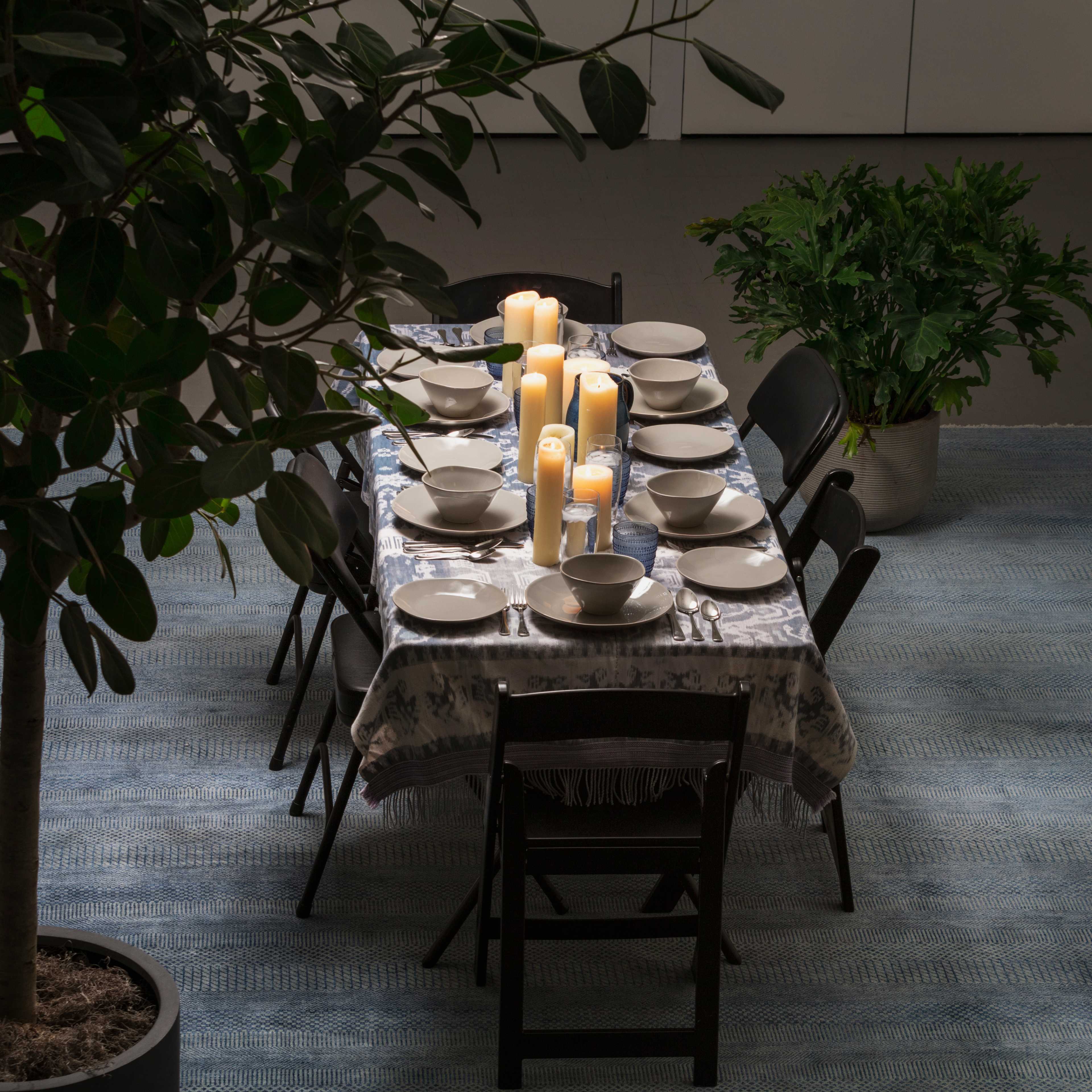 A long dining table is set with plates and candles, surrounded by black chairs, and flanked by indoor plants.