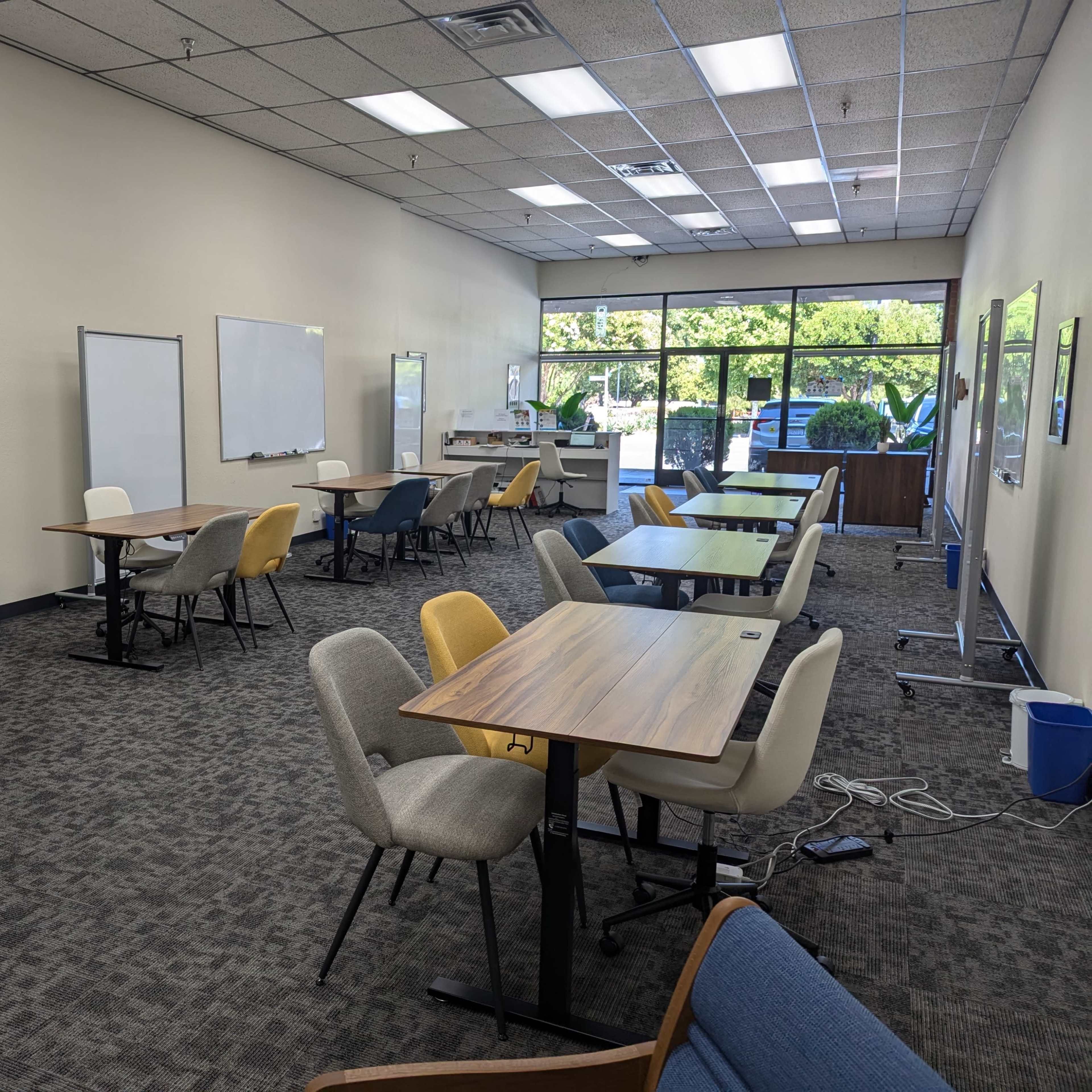 A spacious classroom or meeting room with several empty tables and chairs, whiteboards on the walls, and large windows providing natural light.