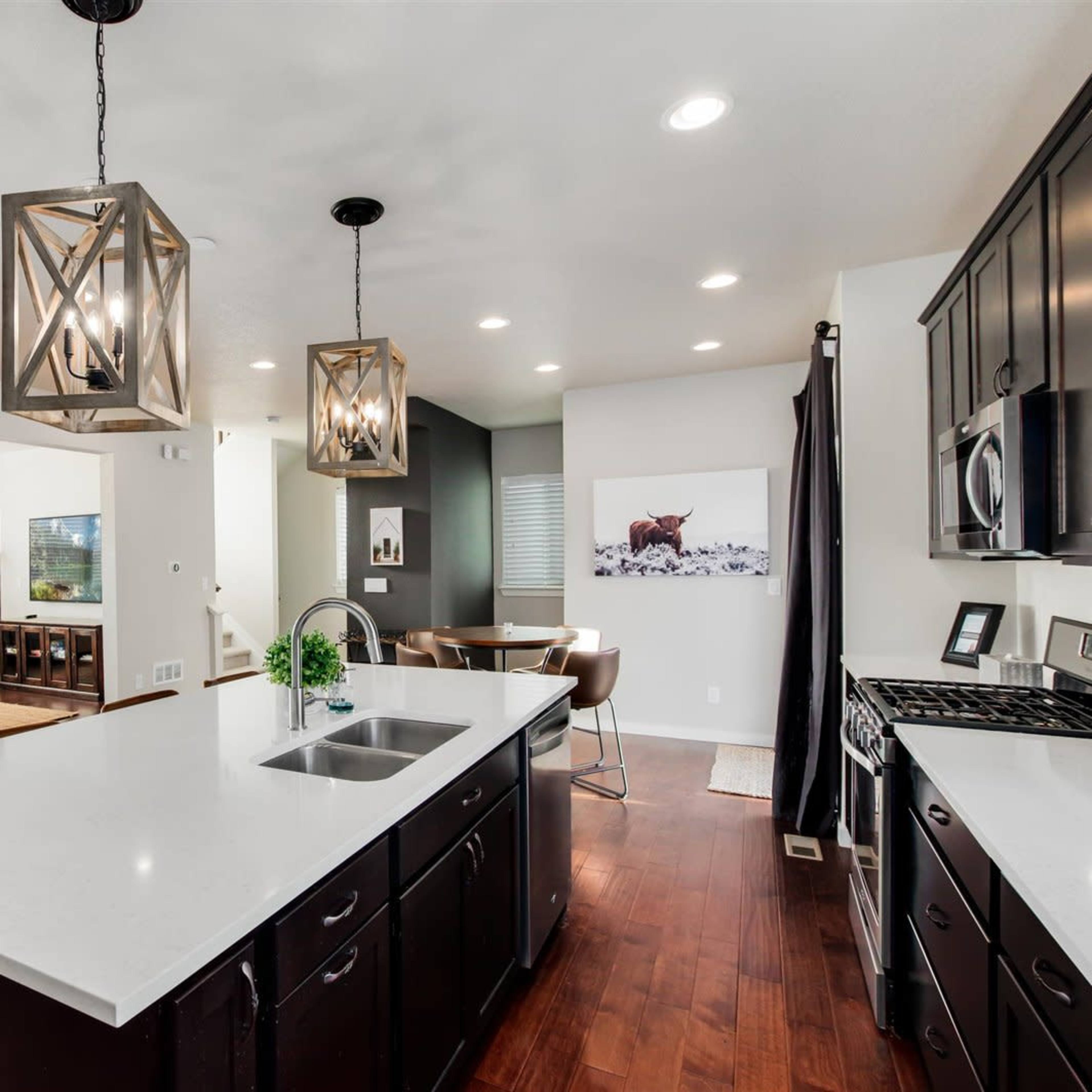 A modern kitchen with dark cabinetry, a white countertop, pendant lighting, and an open layout connecting to a living area with a beige sofa.