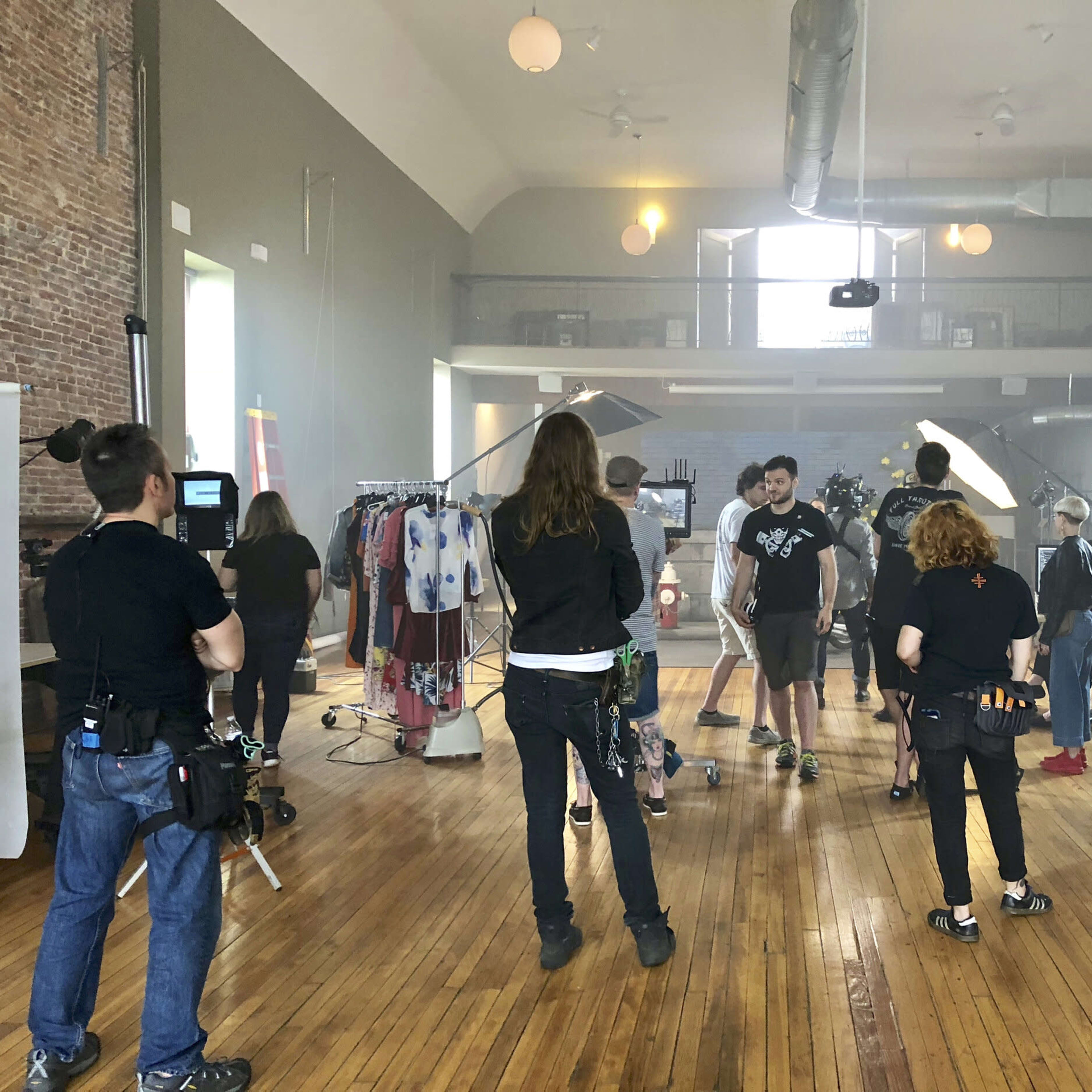 A group of people are preparing for a photoshoot in a spacious, well-lit studio filled with clothing racks and various equipment.