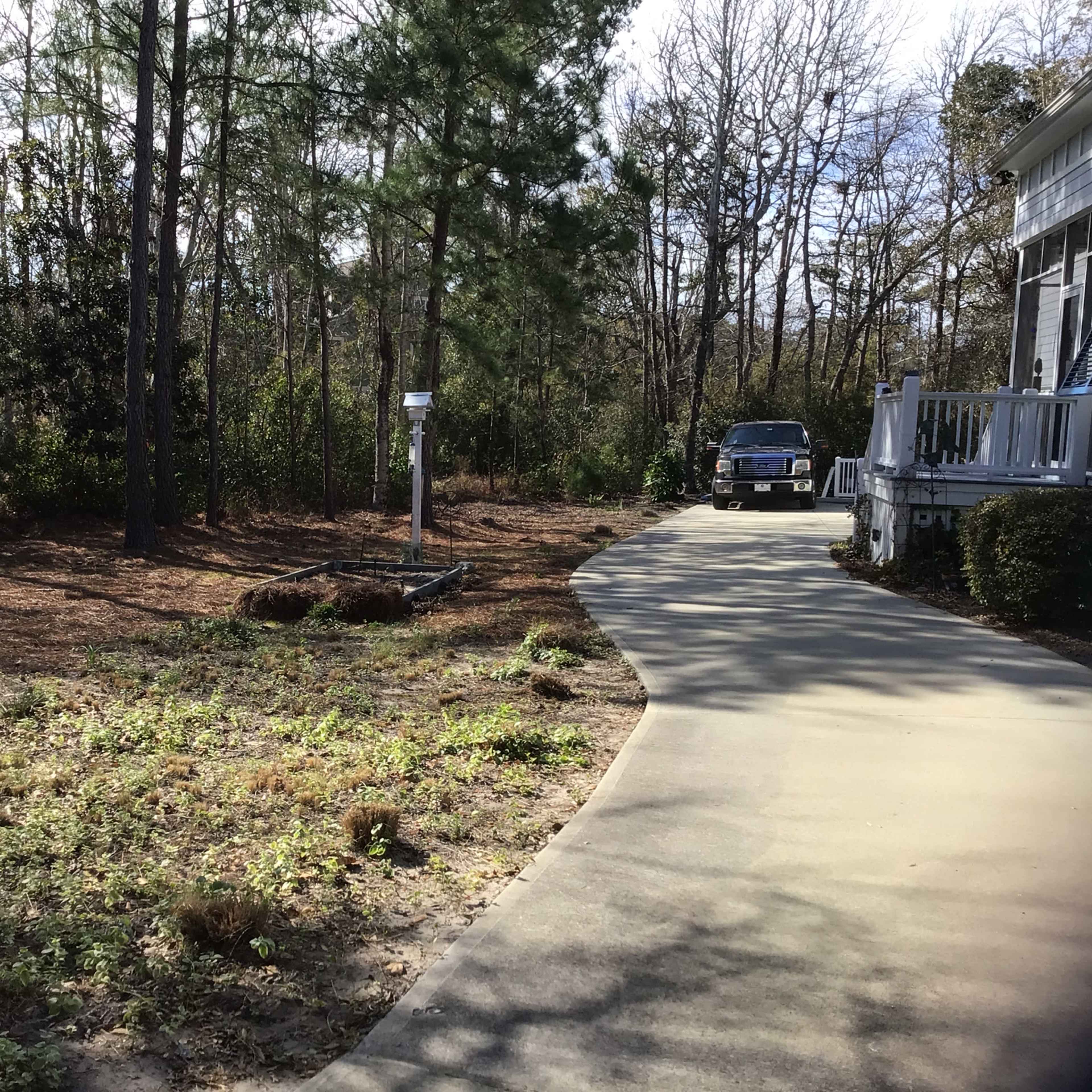 A paved driveway leads past a sparse garden area to a house surrounded by trees.