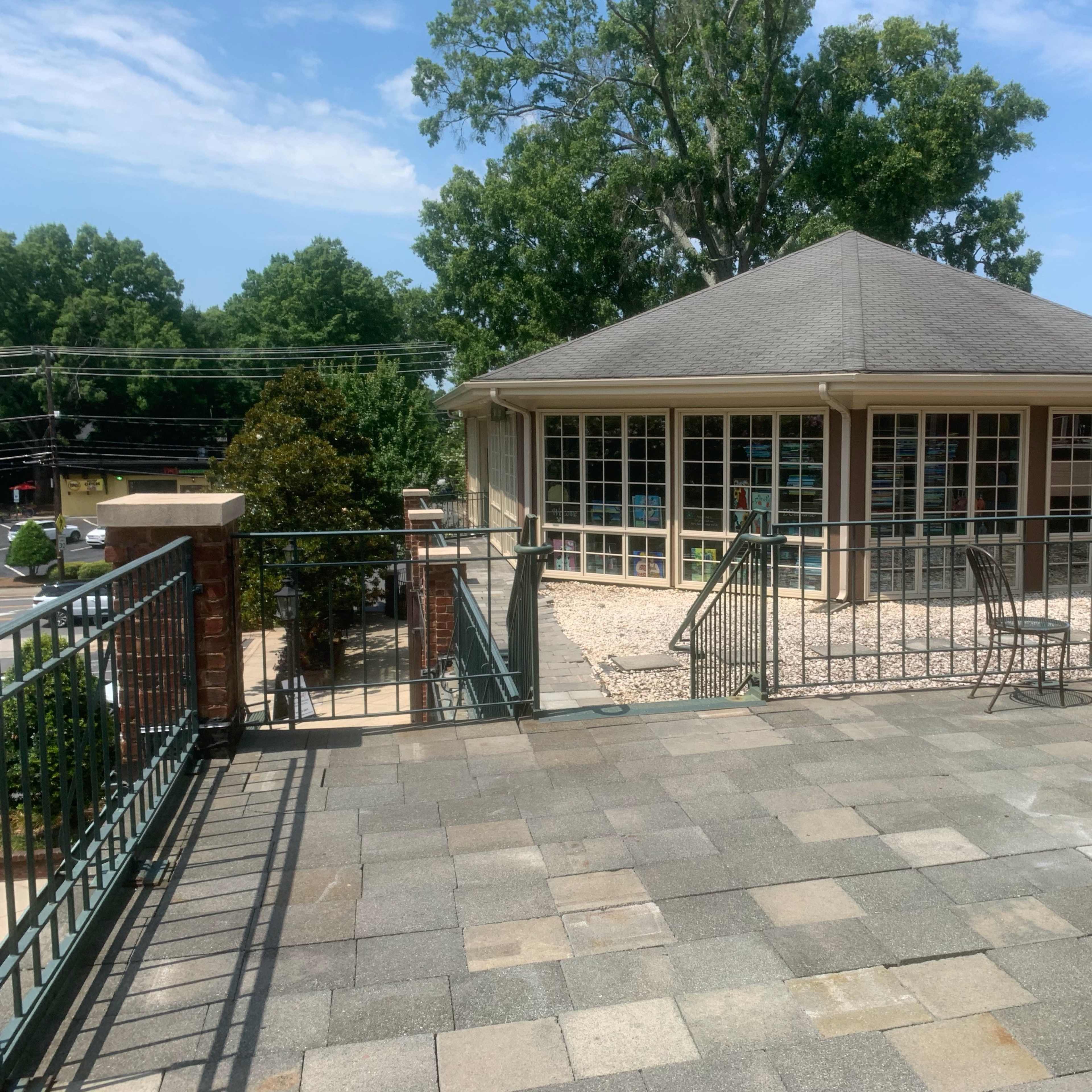 A stone patio extends from a building with large windows and a sloped roof, leading to a landscaped area surrounded by trees and utility lines.