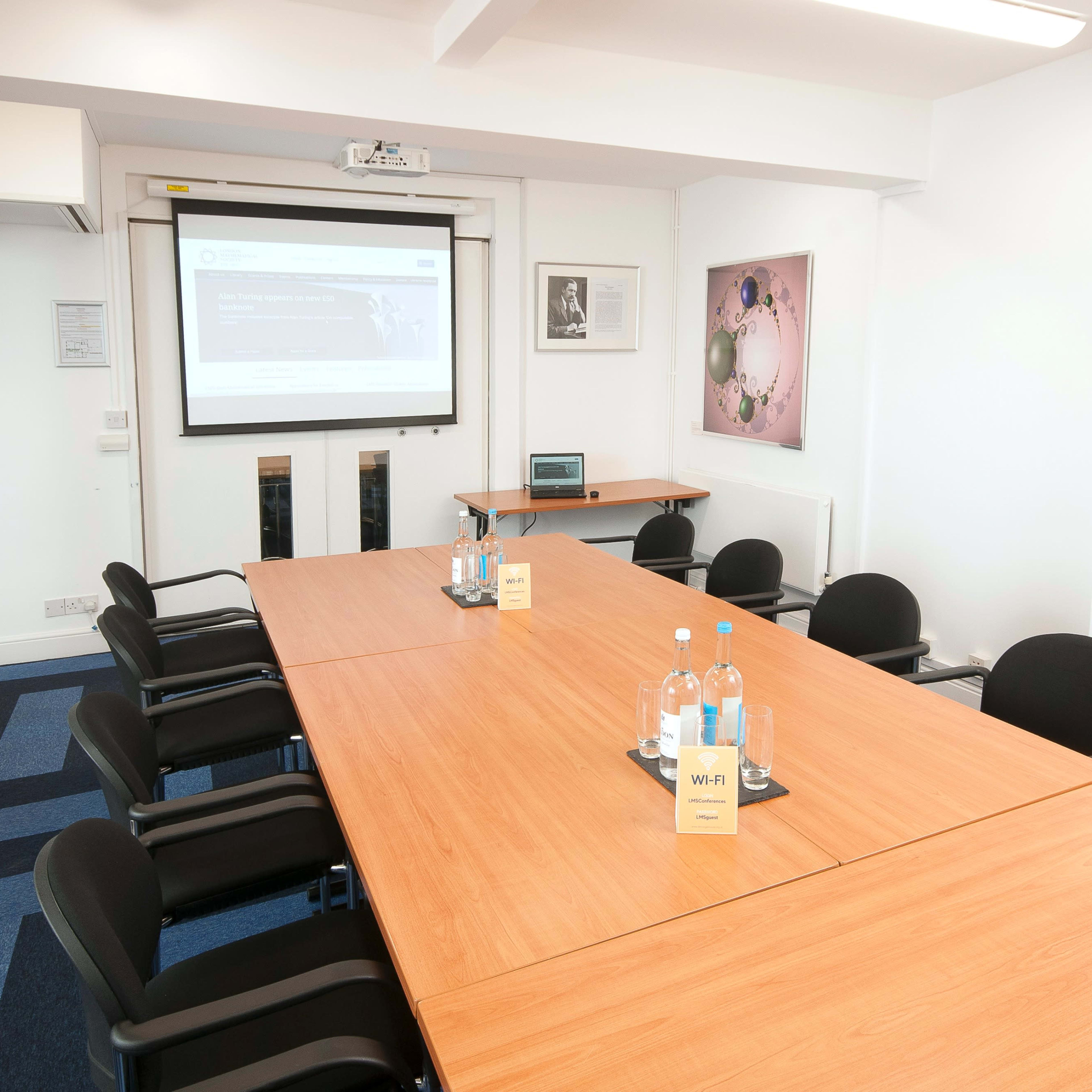 A conference room features a long wooden table surrounded by black chairs, a projector screen mounted on the wall, and bottles of water on the table.