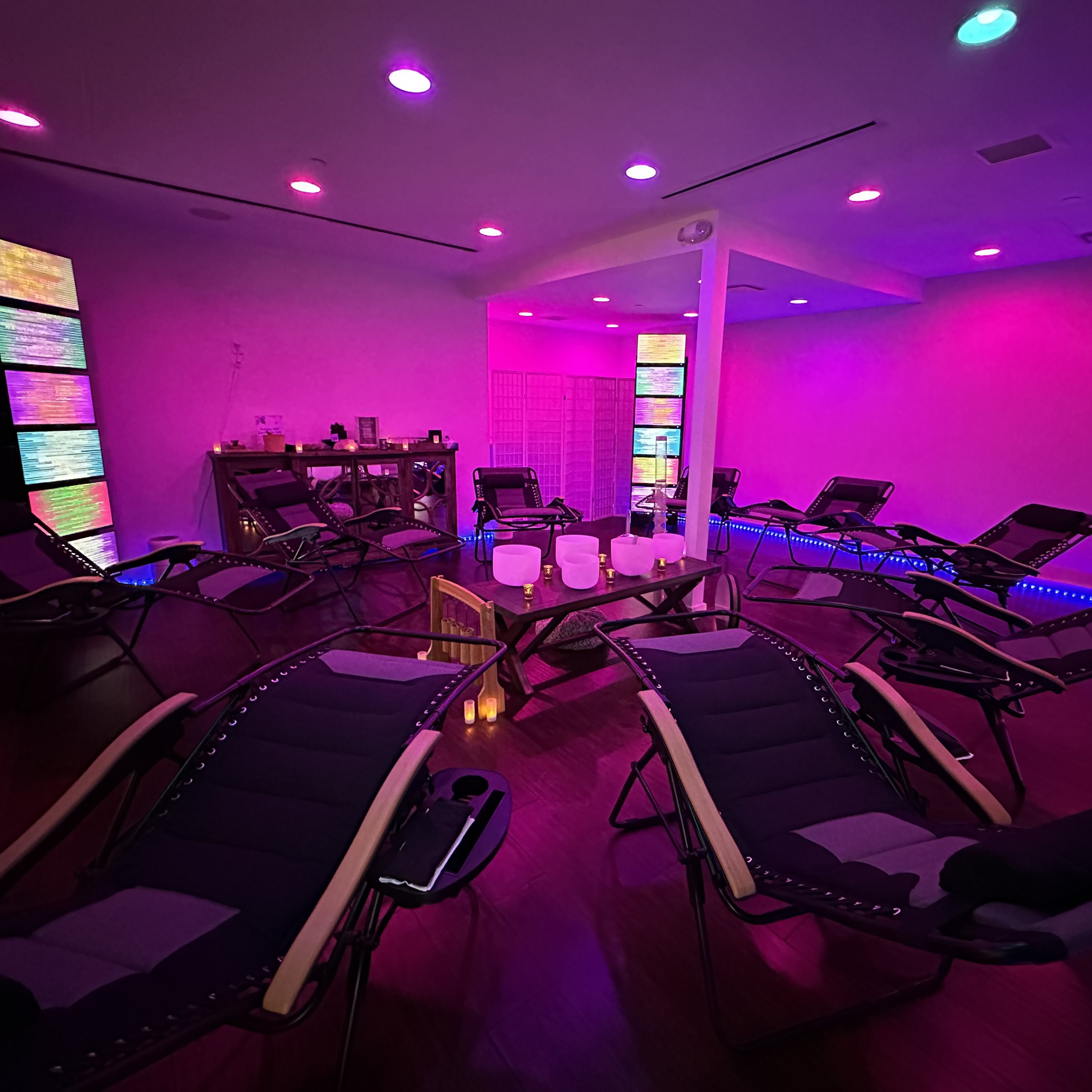 A dimly lit room arranged with reclining chairs in a circular formation, surrounded by colorful ambient lighting and a central table with various objects.
