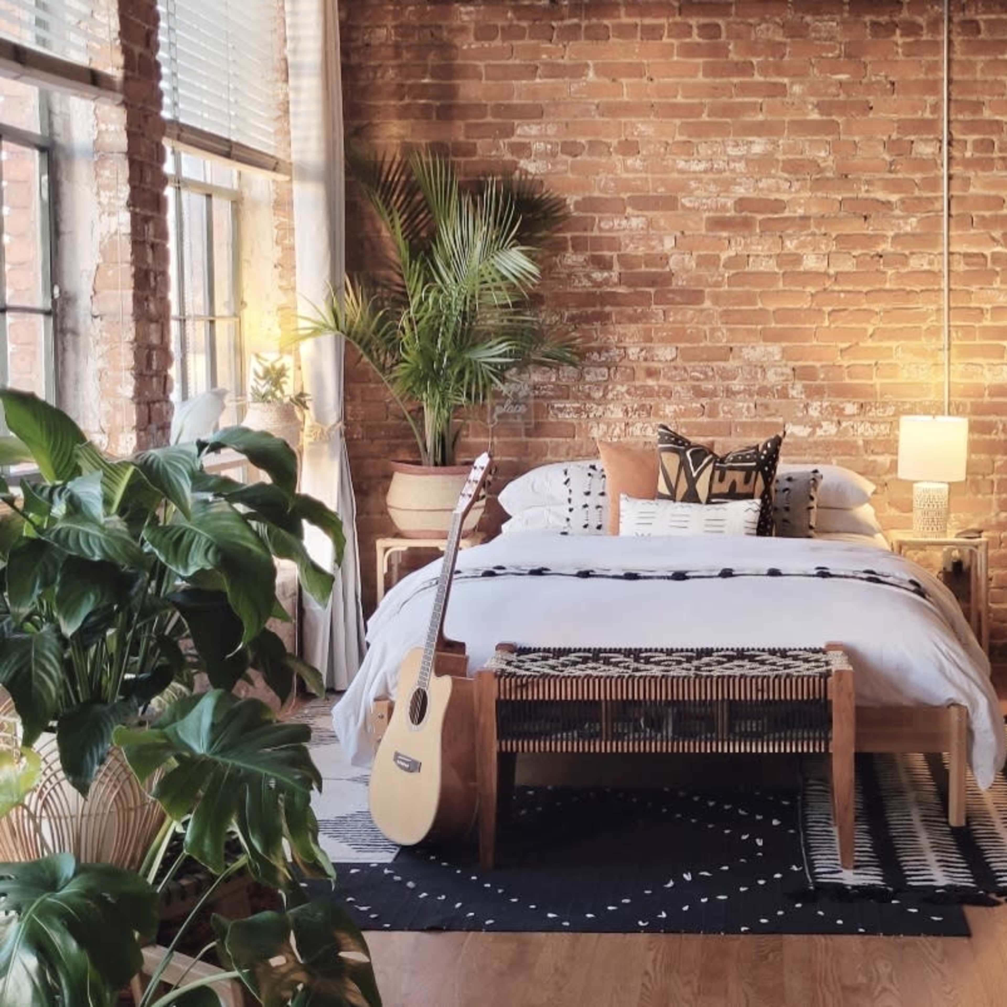 A cozy bedroom with a brick wall, a bed adorned with decorative pillows, and a guitar and plants arranged around the space.
